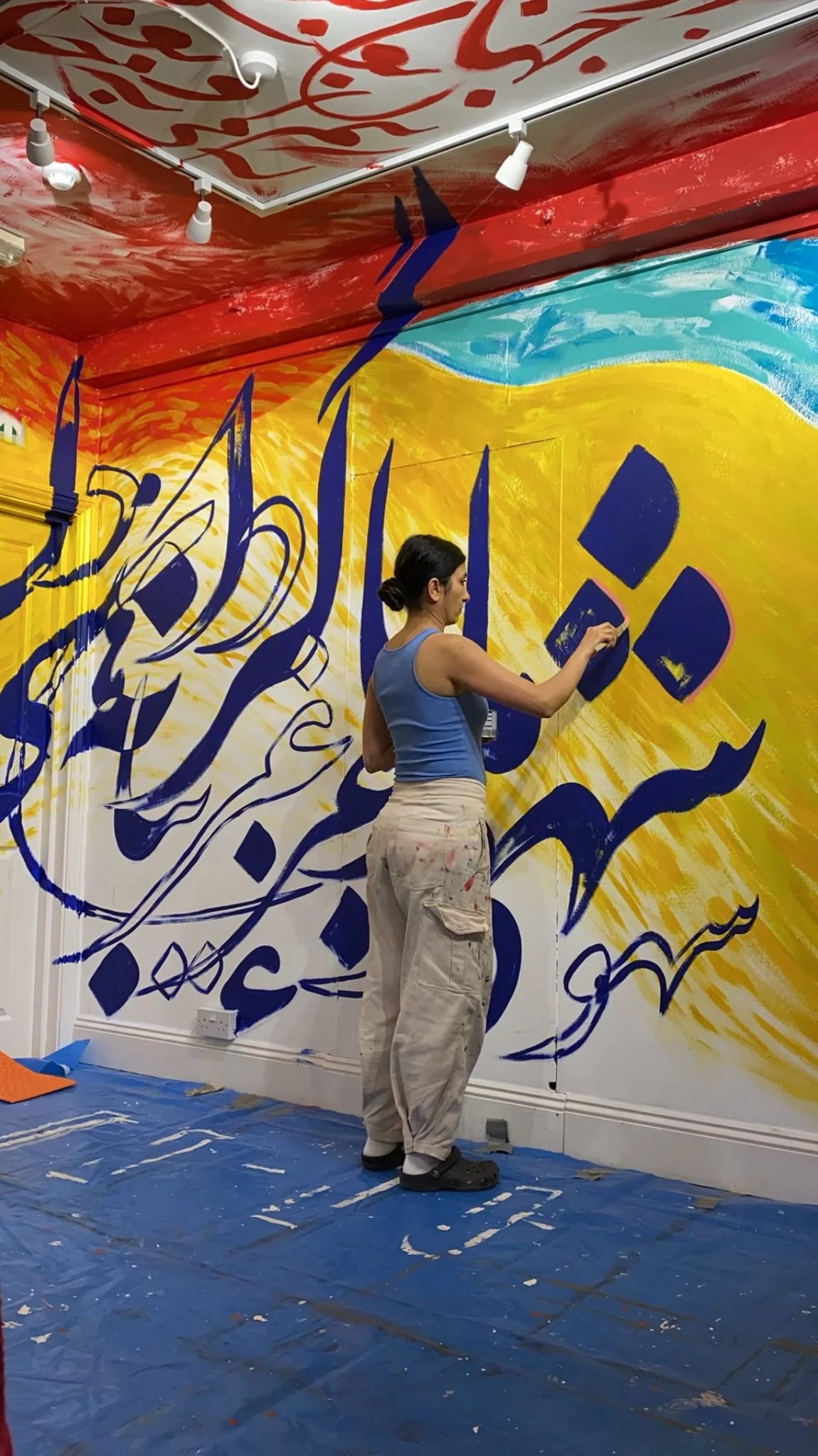 Sahar standing to paint black Persian calligraphy detail on a gold, green, and red wall.jpeg