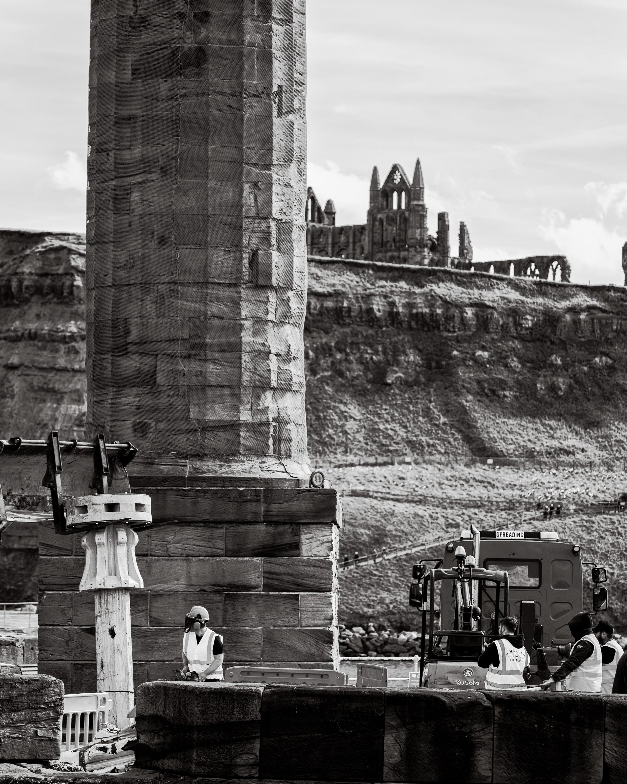 An excavator lifts a handcrafted timber capstan into place on the harbour at Whitby with the silhouette of the famous Whitby Abbey in the background.