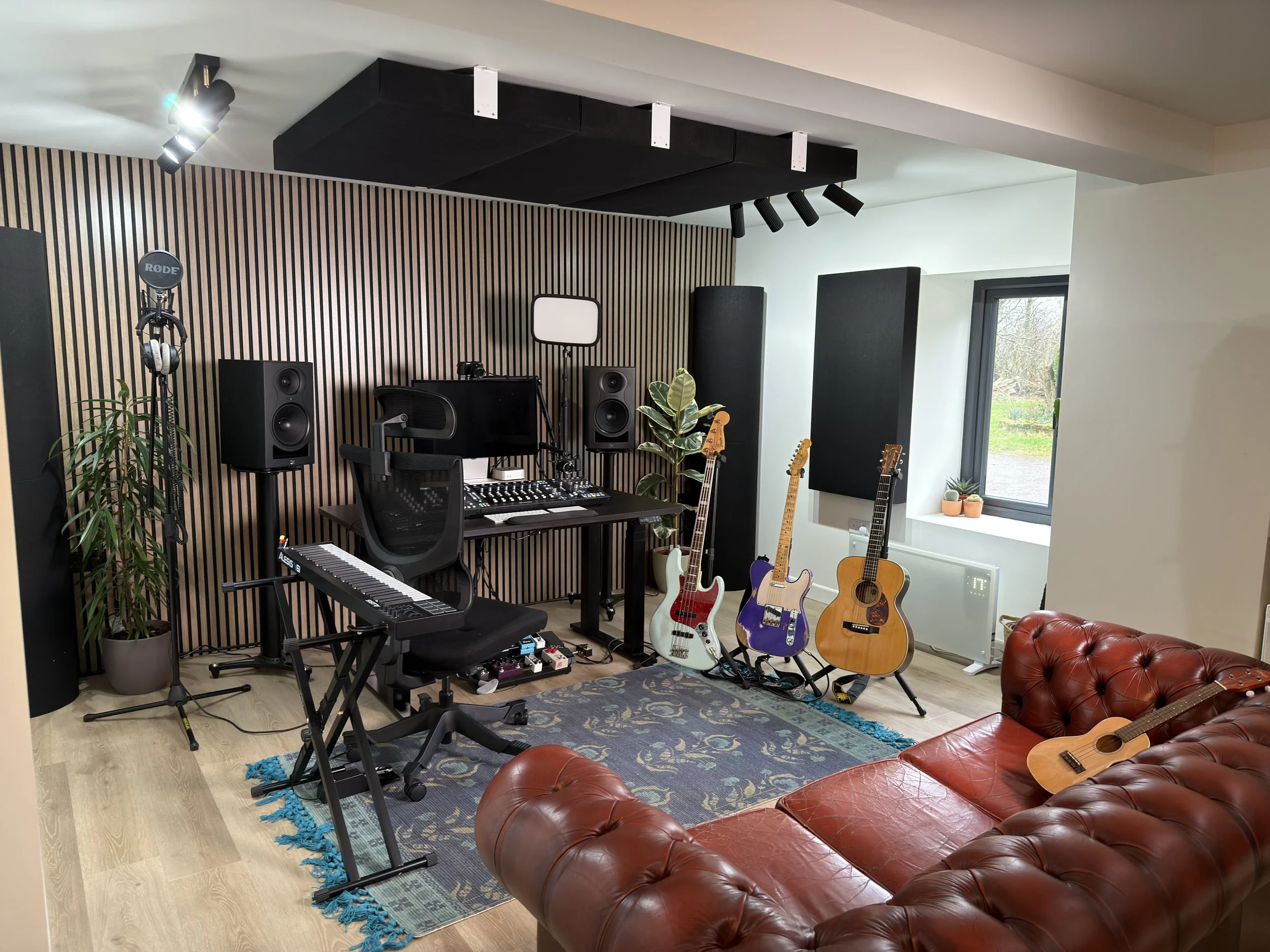 A music studio with a red leather couch, multiple electric and acoustic guitars, a keyboard, and a mixing console, all set against a black wooden wall with soundproofing panels and a window showing an outdoor scene.