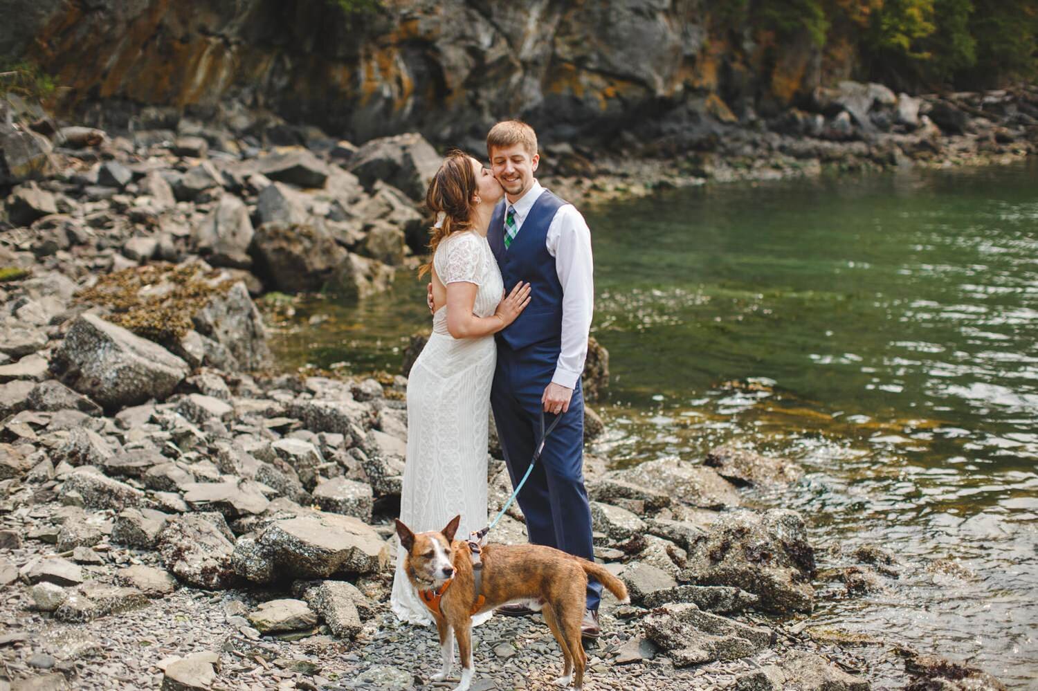  bride kisses groom on the cheek in an art deco mid century wedding dress. Groom in blue vest and tie, holding dogs leash on a rocky beach 