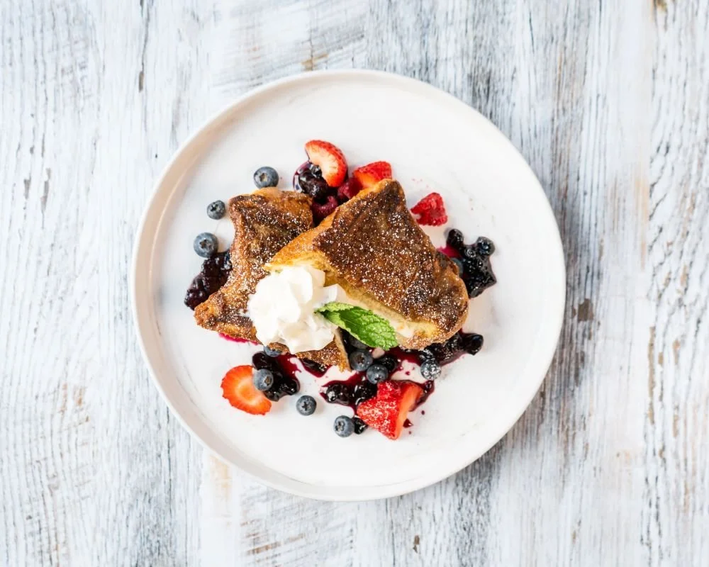 French toast topped with whipped cream, mint, strawberries, blueberries, and berry sauce on a white plate