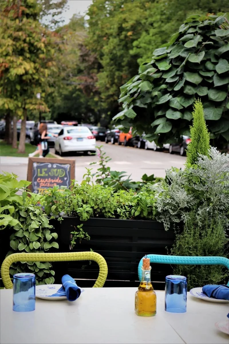 Table set for outdoor dining with two blue glasses, napkins, and a bottle of oil, surrounded by green plants and trees in a street-side patio, with parked cars and a woman walking in the background.