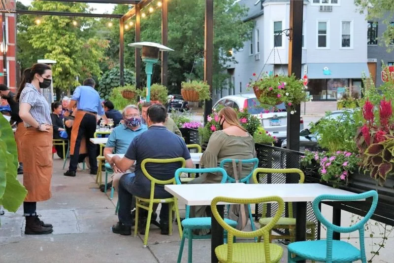 Outdoor restaurant patio with several seated patrons and waitstaff, decorated with hanging flower baskets and string lights.