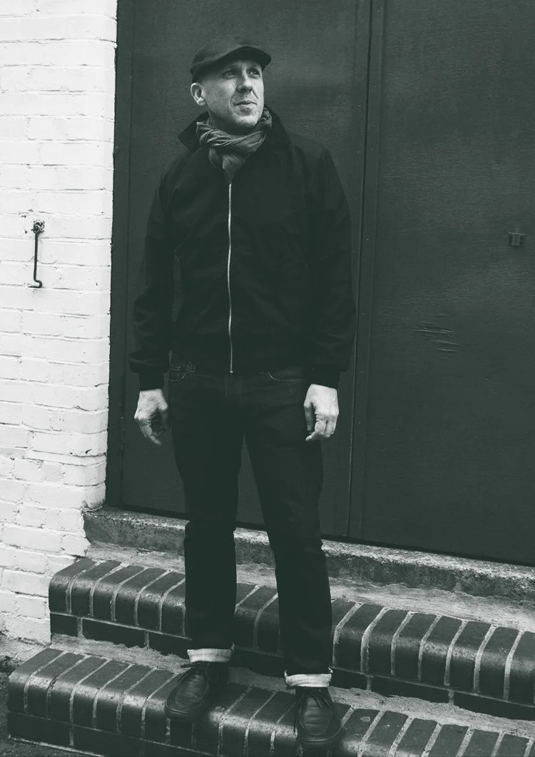 A man standing on brick steps in front of a dark wall, wearing a hat, scarf, black jacket, jeans, and shoes, looking to the side.