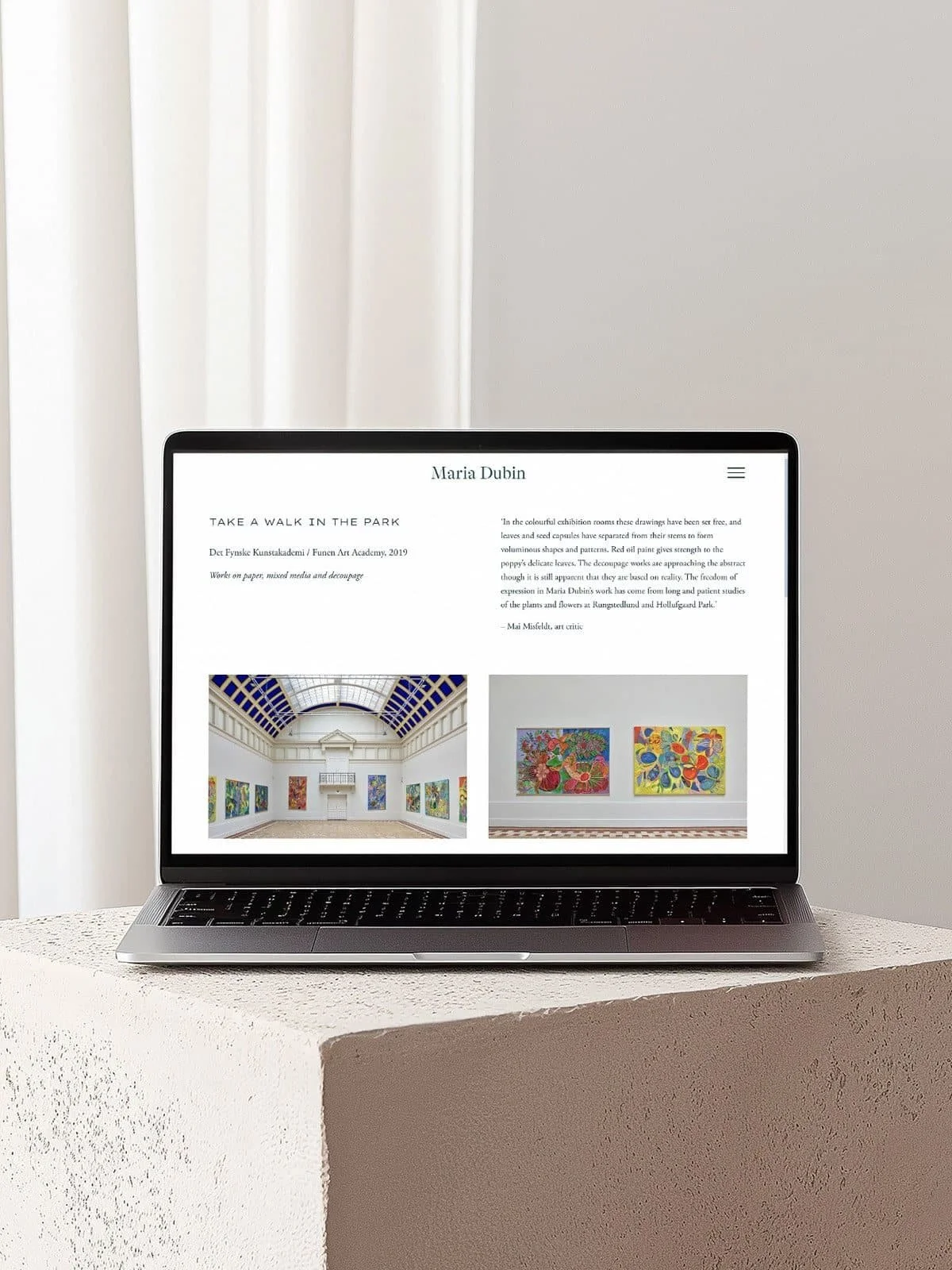 Open laptop displaying an artist website with colorful paintings in a gallery setting, placed on a textured white surface in front of white curtains.