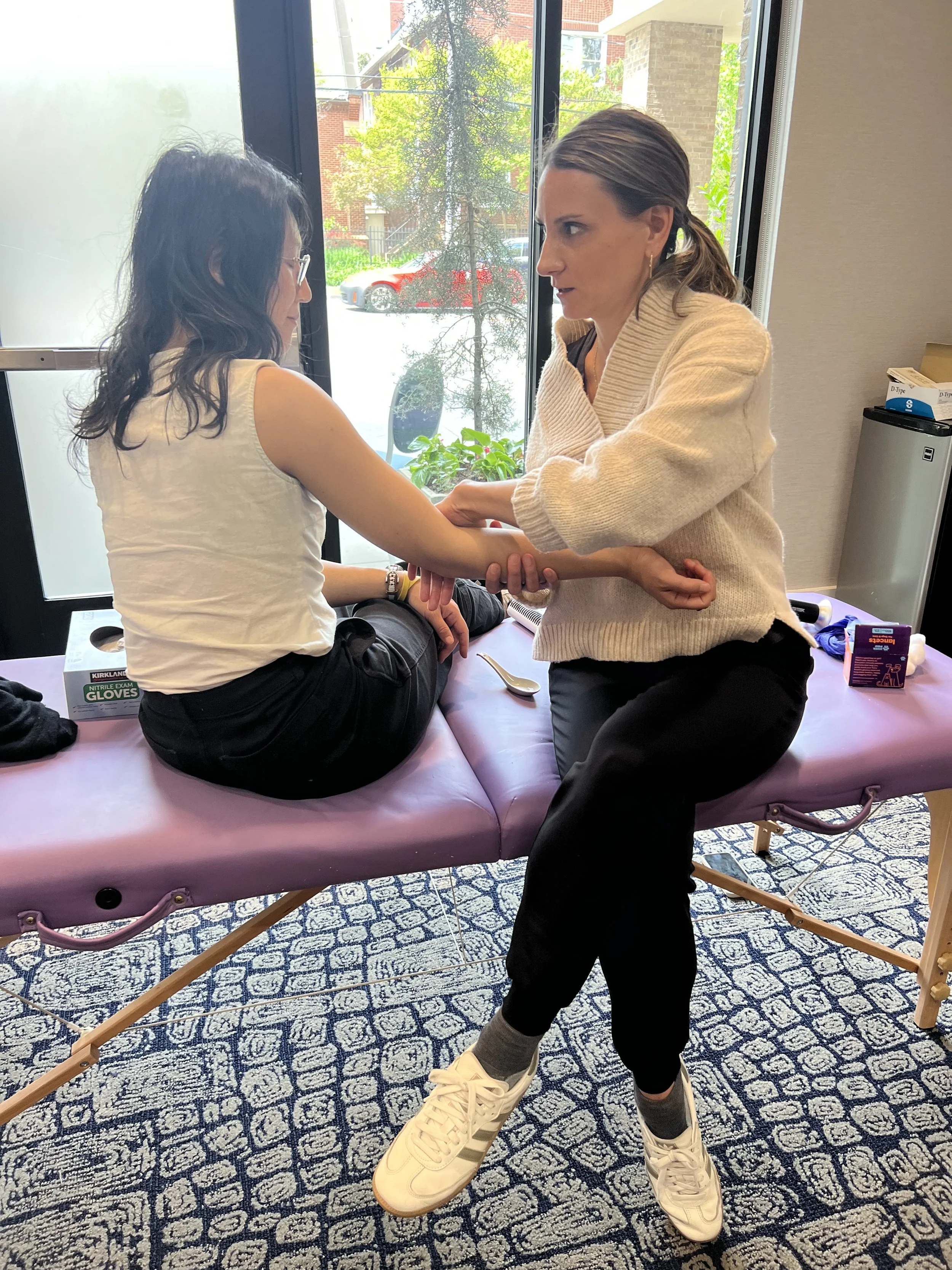Two women are sitting on a purple exam table, practicing Chinese medicine dynamic pulse taking in MG's post graduate practical class.