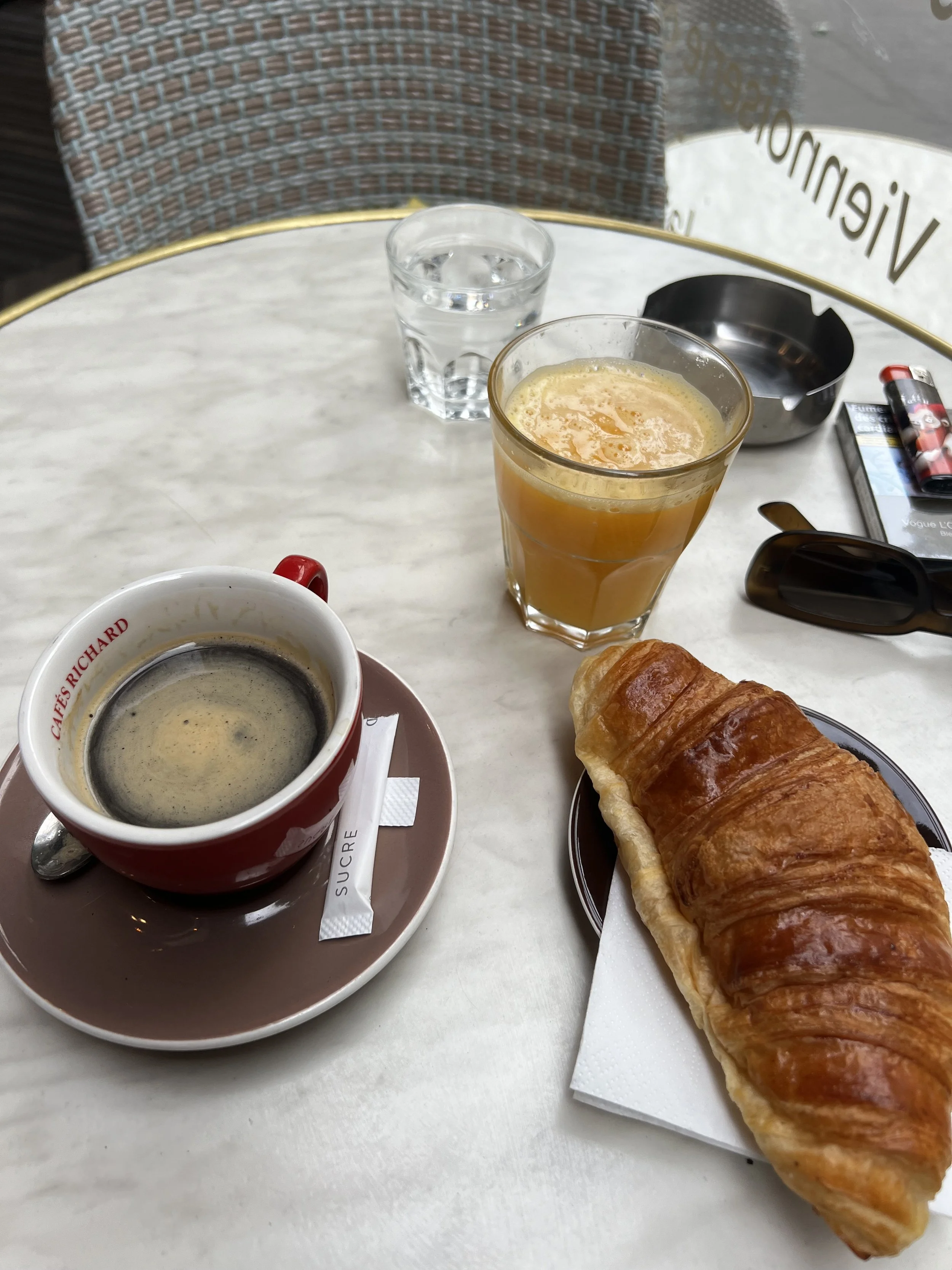 A breakfast scene on a white marble table with a cup of coffee, a glass of orange juice, a glass of water, a croissant on a plate, sunglasses, a cigarette pack, and a lighter.