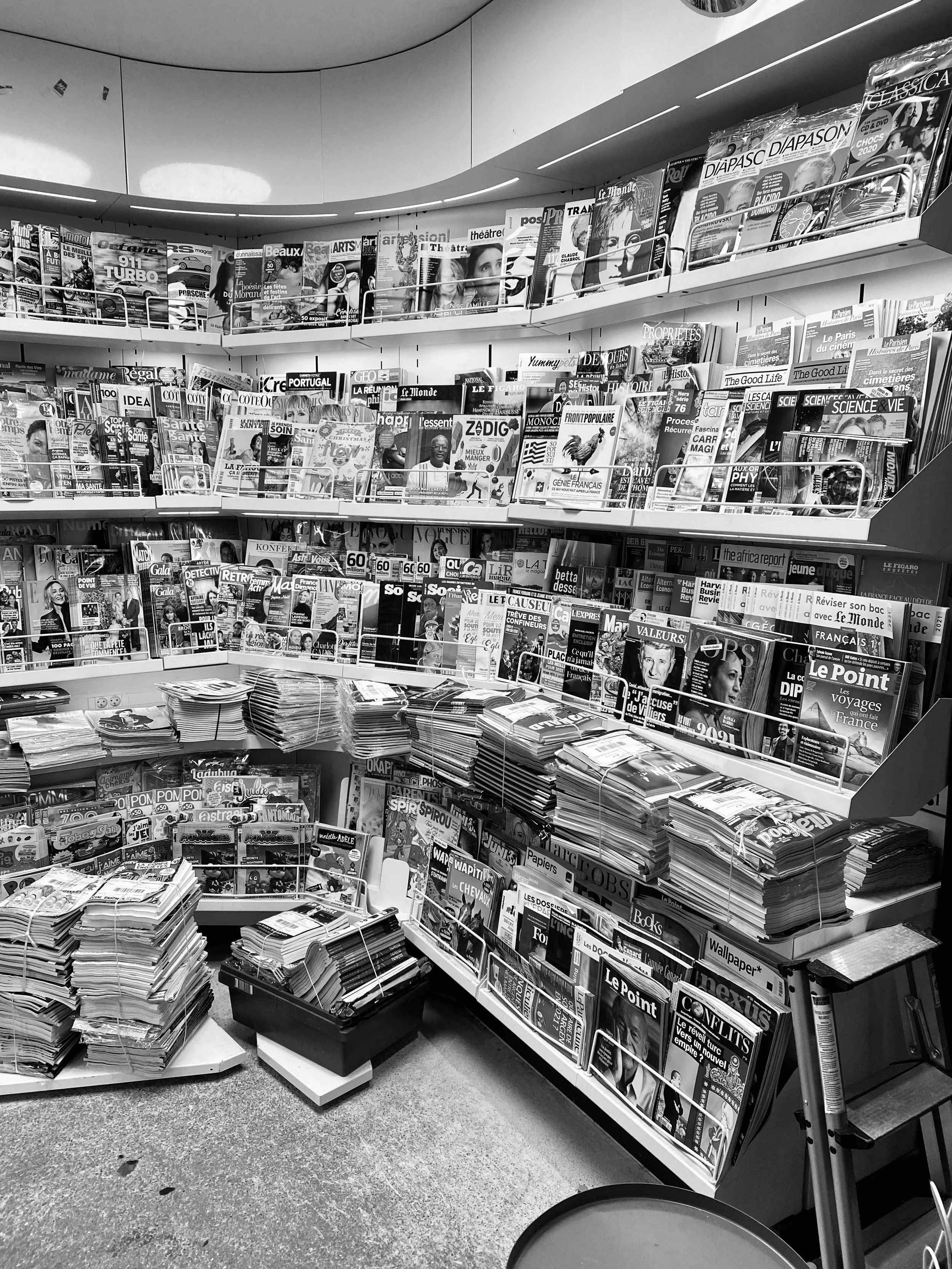 A black and white photo of a magazine stand with multiple shelves filled with various magazines and newspapers.