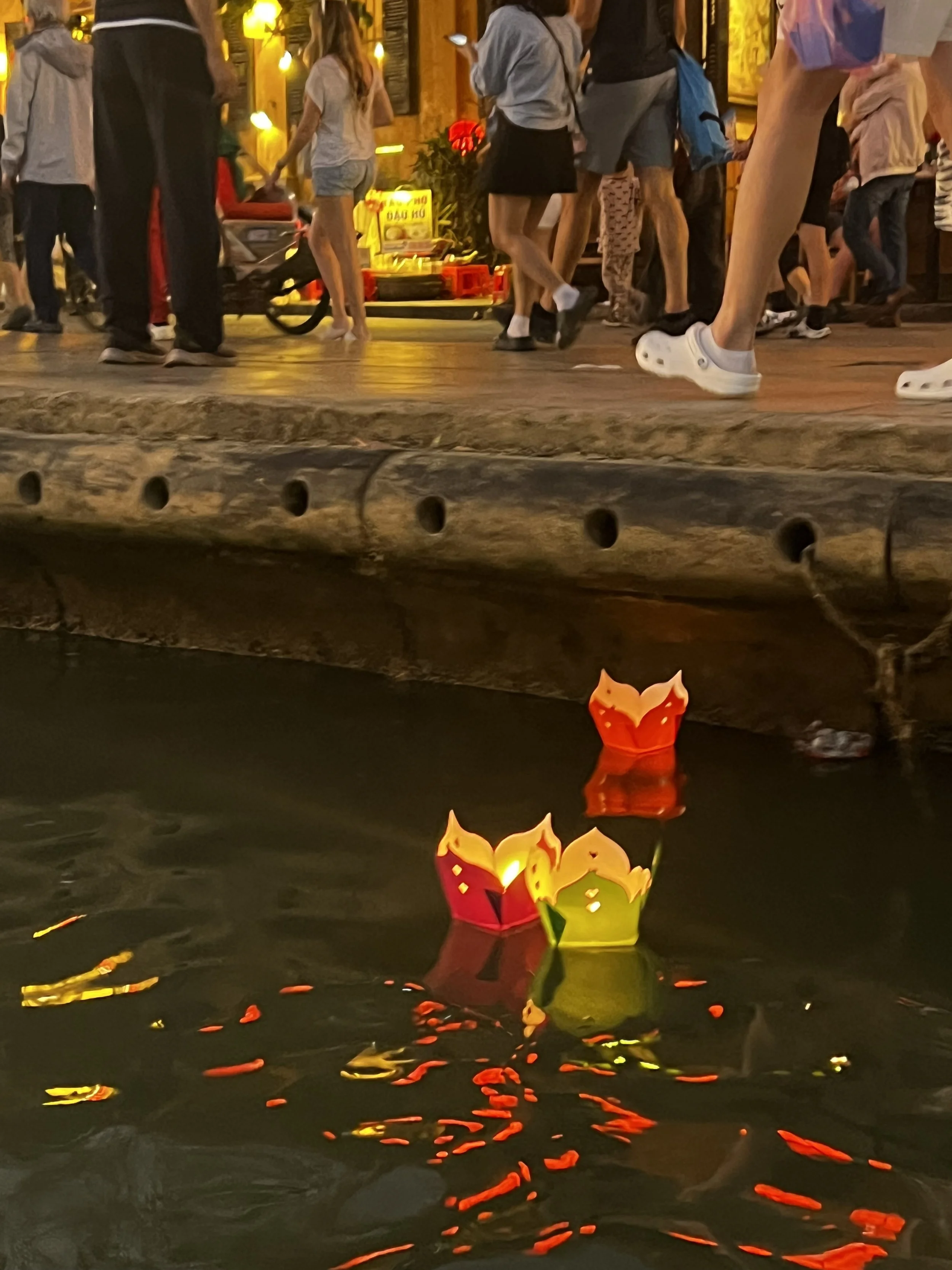 People walking along a busy sidewalk at night near a body of water with floating lanterns, some lit with candles, reflecting on the water's surface.