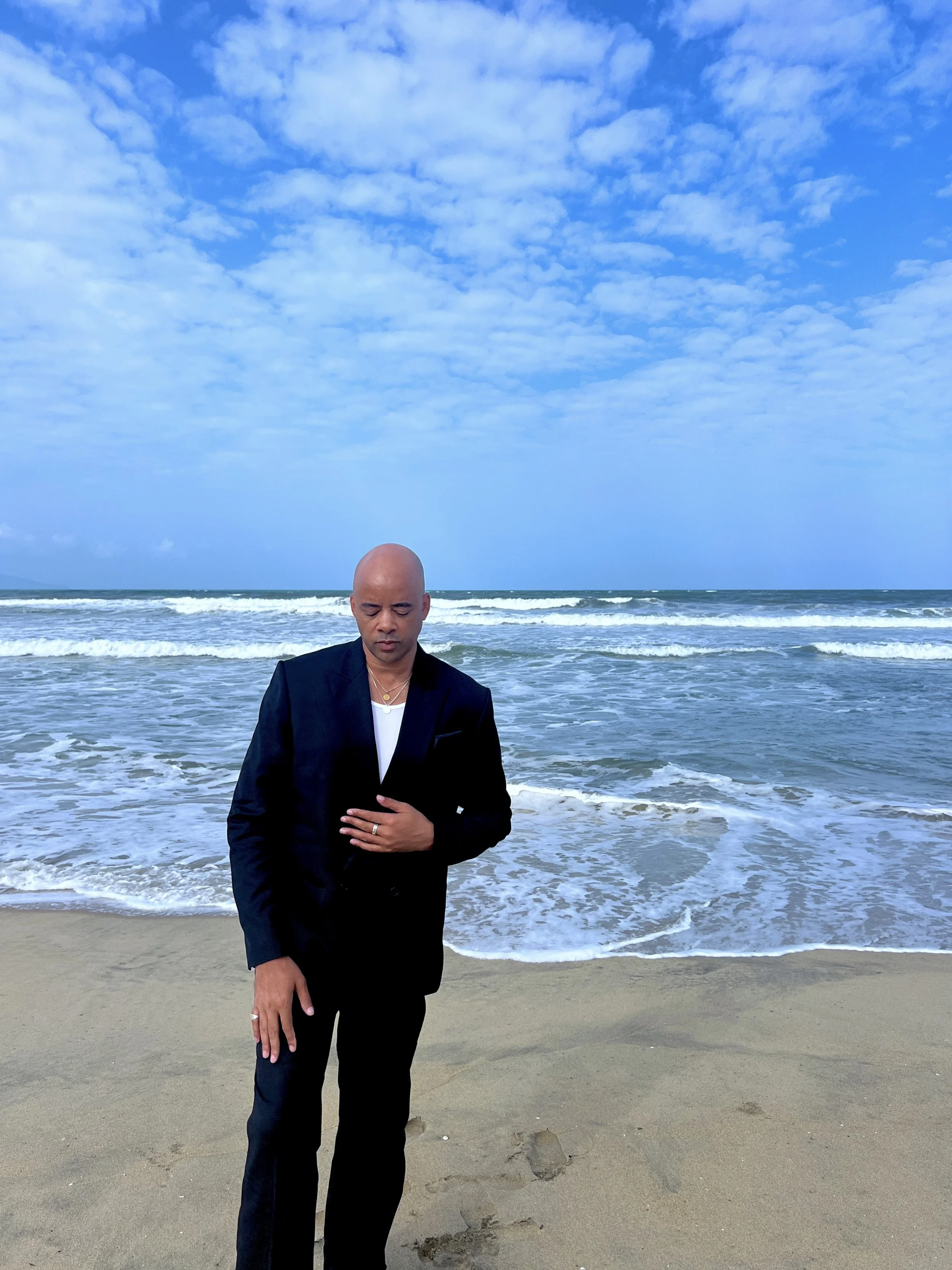 Ethan Ward stands on a sandy beach in a black suit and white shirt, looking down with one hand resting on his chest as ocean waves roll in behind him under a bright, cloud-filled blue sky.
