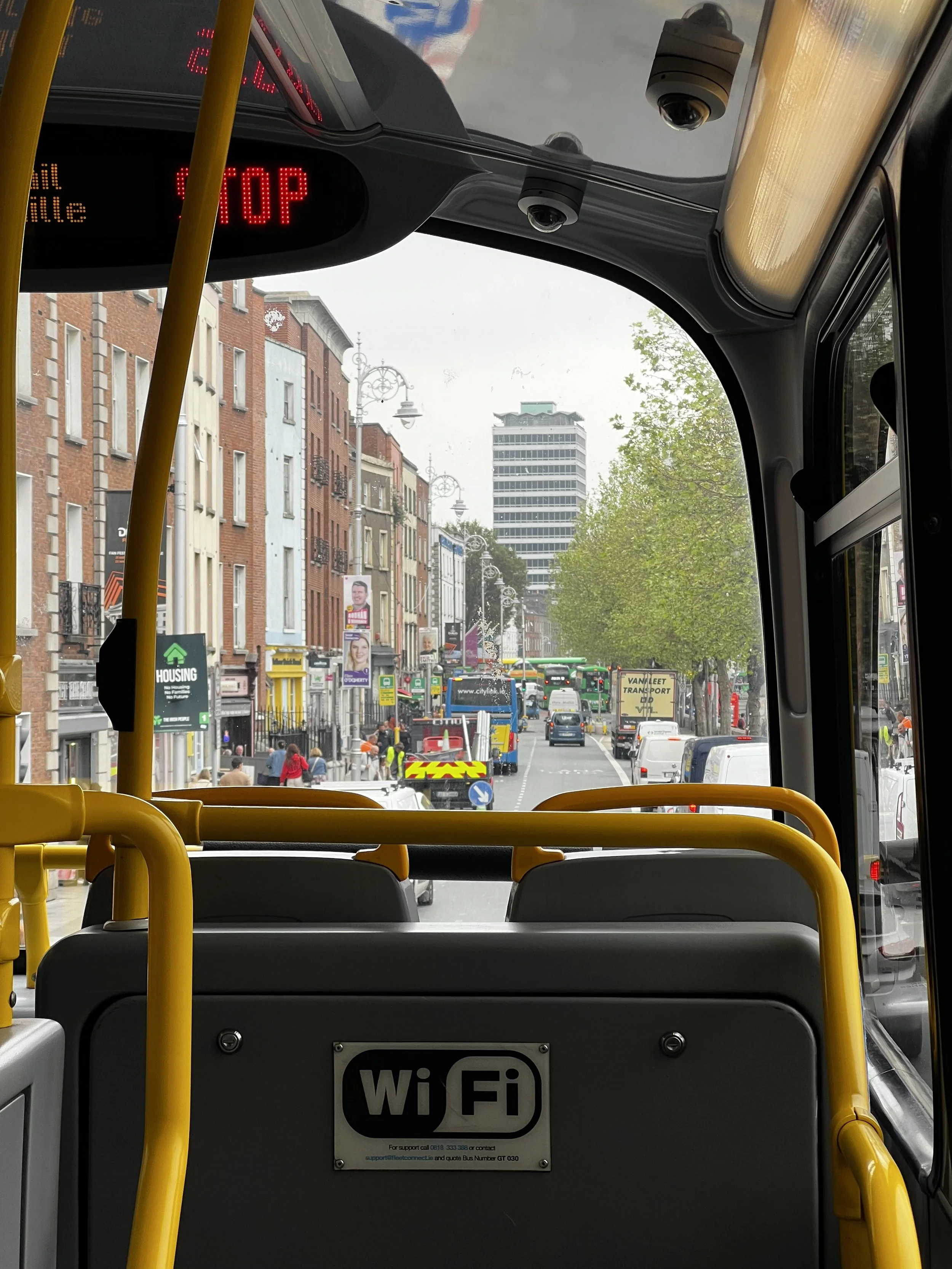 View from inside a bus looking out at a city street with buildings, trees, and traffic. The bus has yellow handrails and displays a Wi-Fi sign on the back.