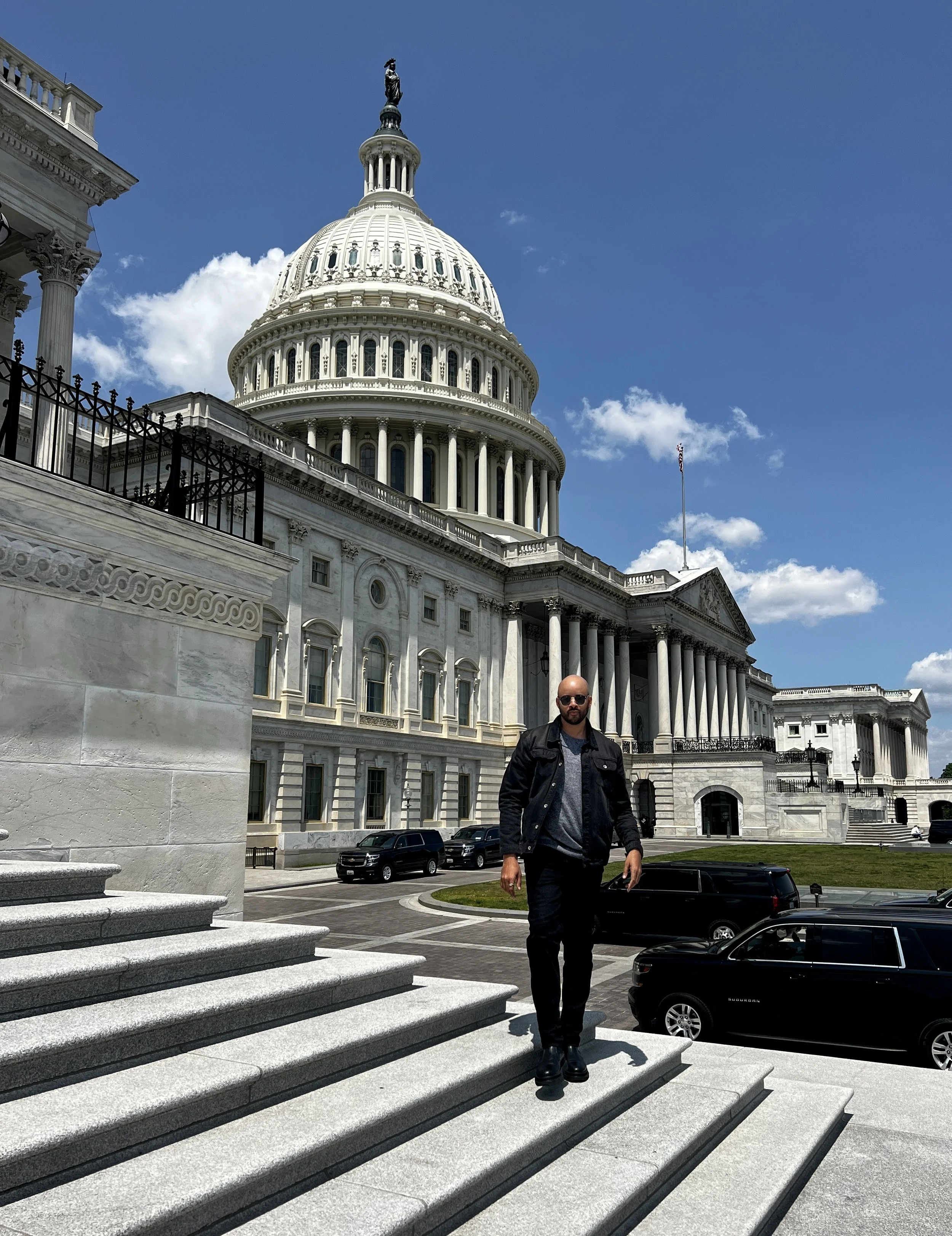ETHAN WARD standing on steps in front of the U.S. Capitol building in Washington, D.C., under a blue sky with clouds.