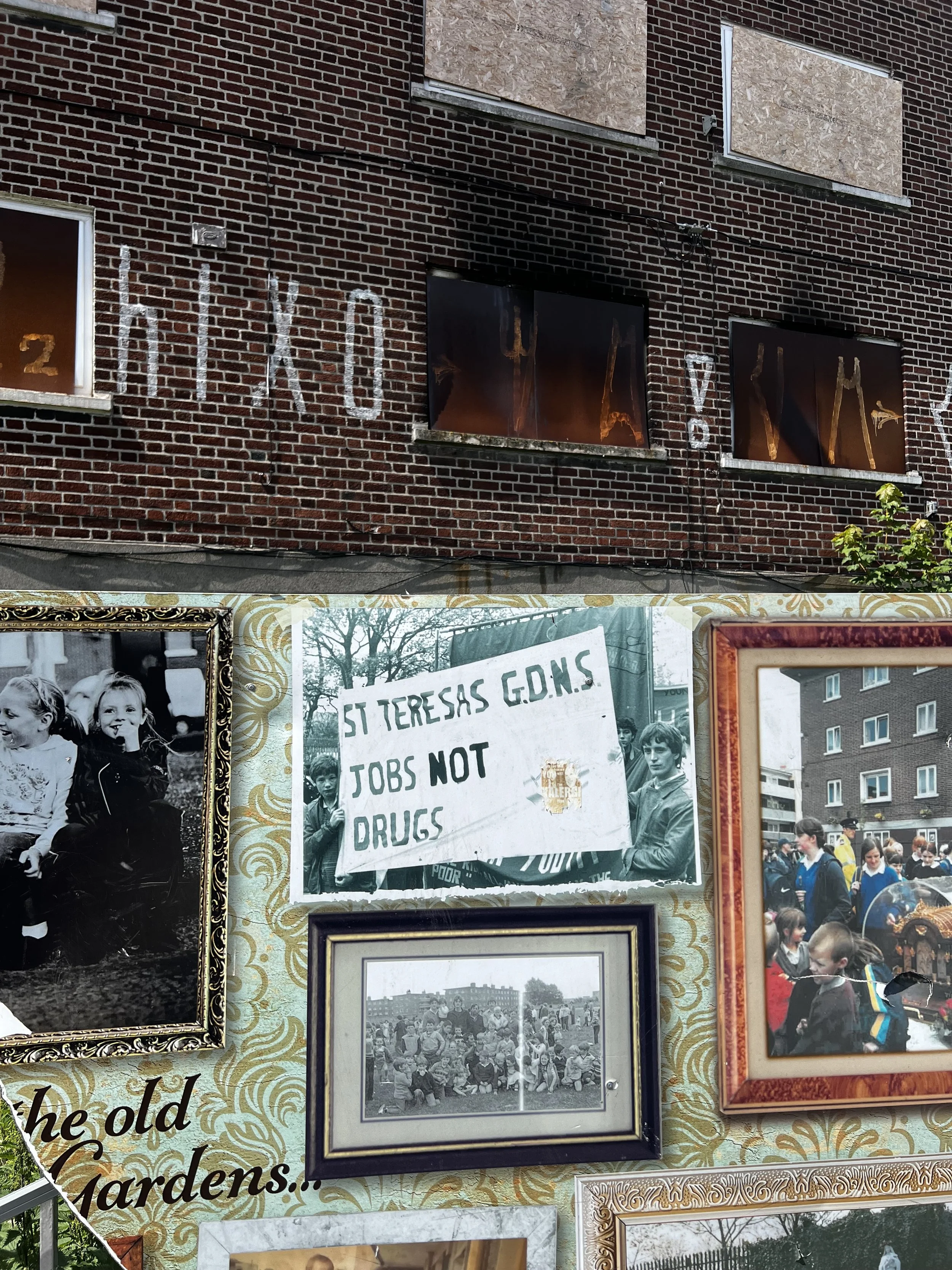 A brick building with boarded-up windows and graffiti, along with a display of framed photos and a protest sign reading 'St Teresas G.D.N.S Jobs Not Drugs'.
