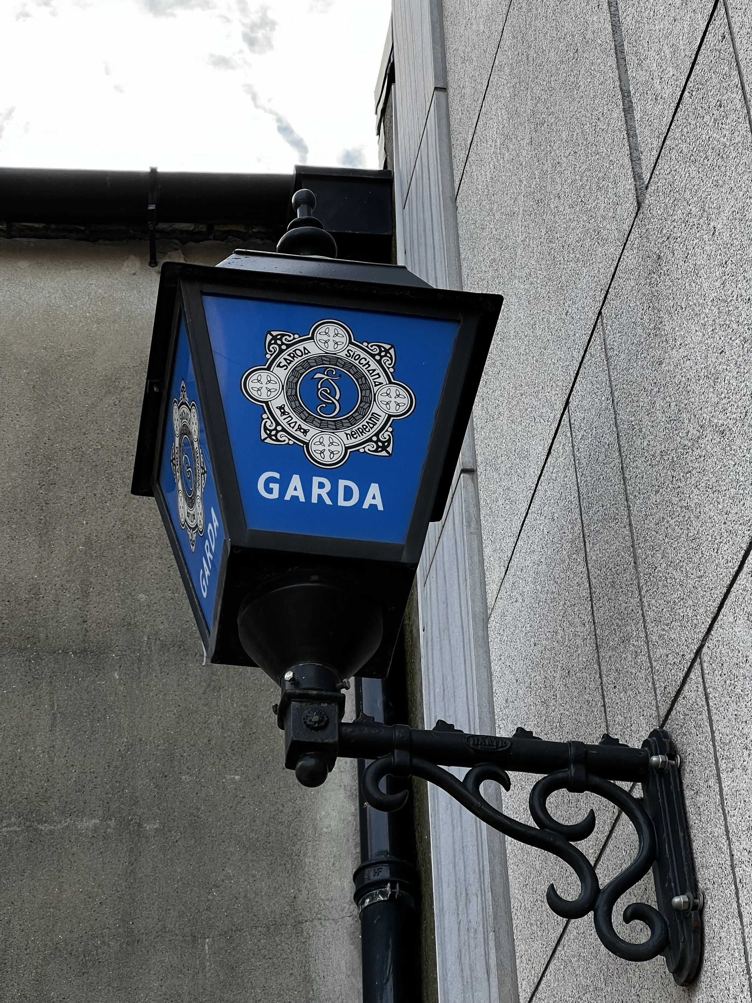 Street lamp with Garda badge and logo, mounted on a decorative black wrought iron bracket attached to a beige stone wall.