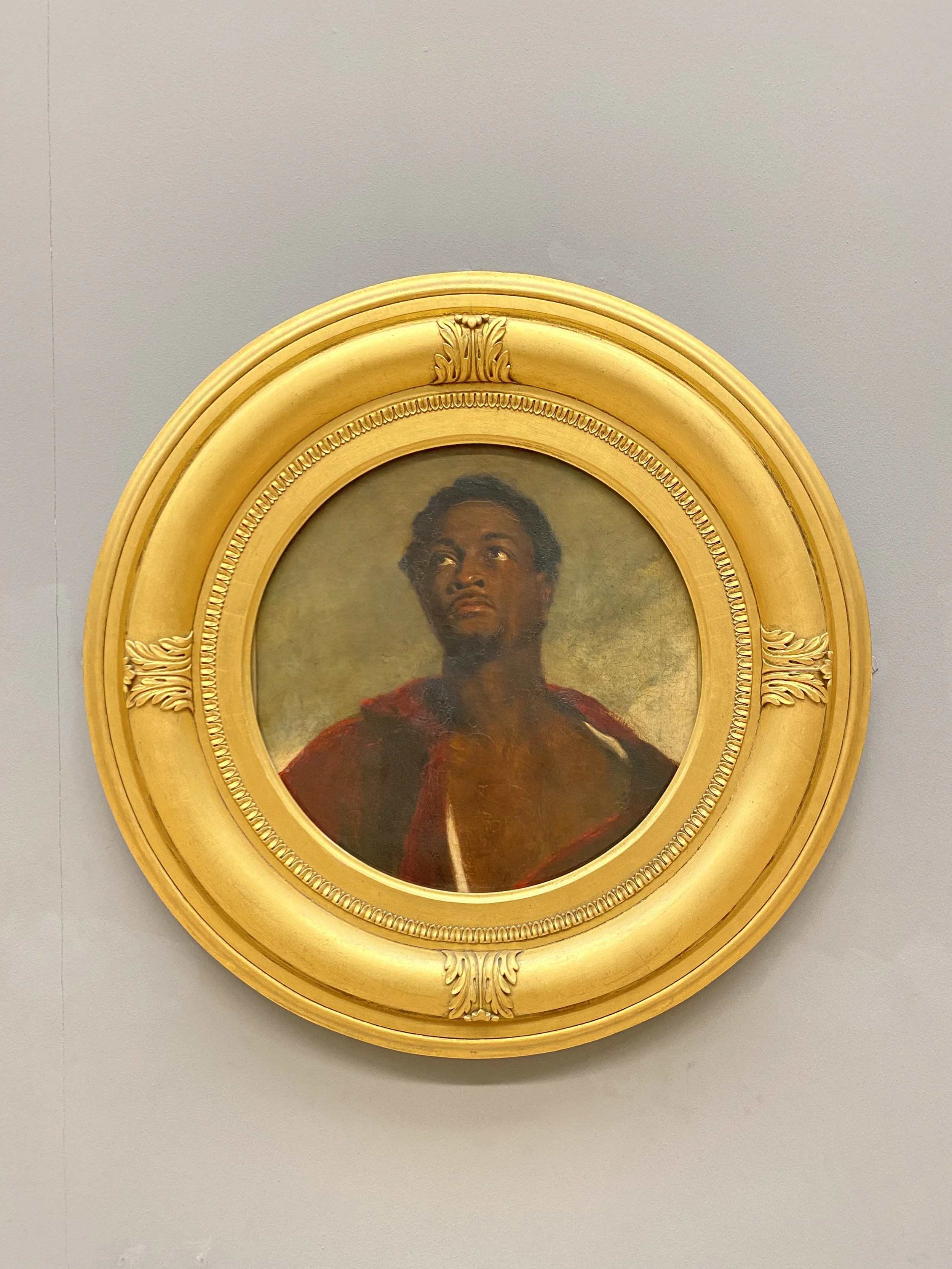 Portrait of a dark-skinned man with dark curly hair, wearing a red and black garment, gazing upward, framed by an ornate gold oval frame, hung on a plain wall.