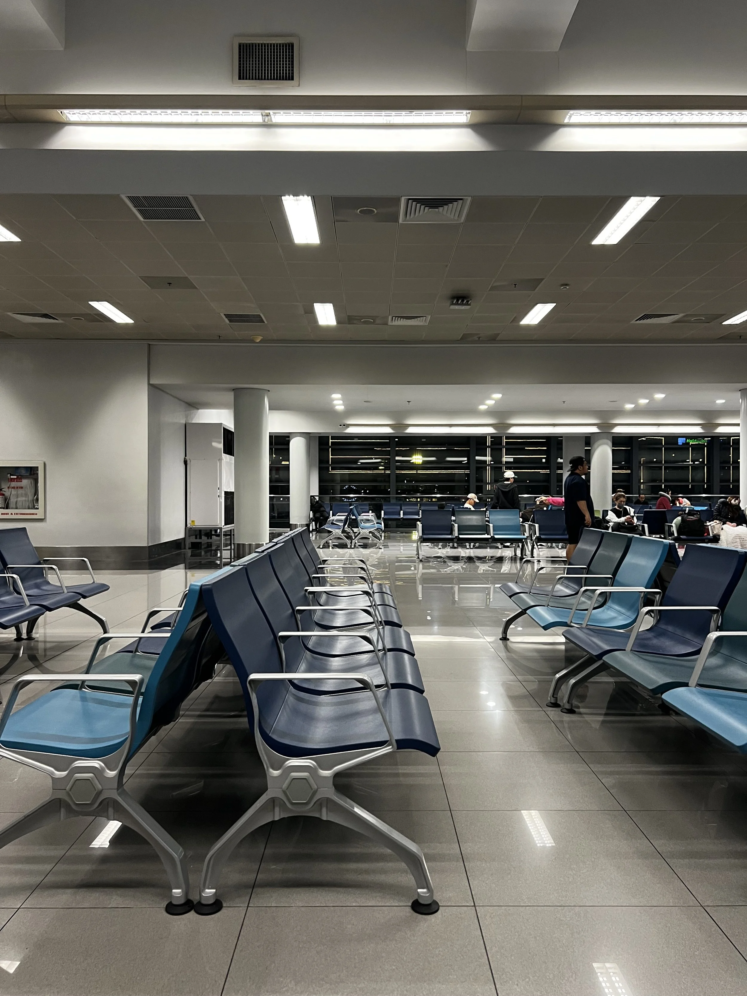 Empty seating area in an airport waiting lounge with a few passengers seated and some standing, with large windows in the background.