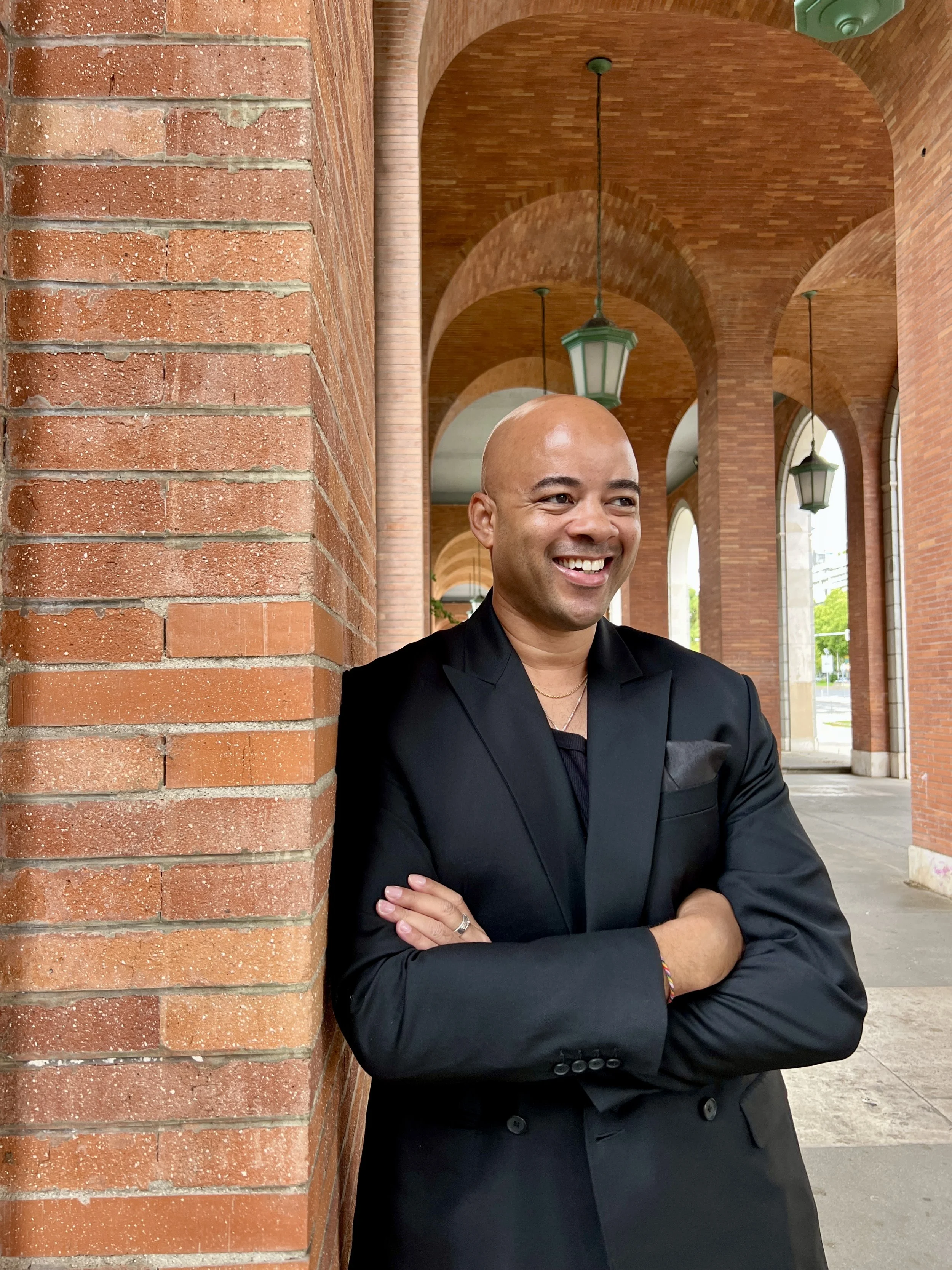 Ethan Ward is smiling in this headshot wearing a black suit with his arms crossed while leaning against a red brick wall.