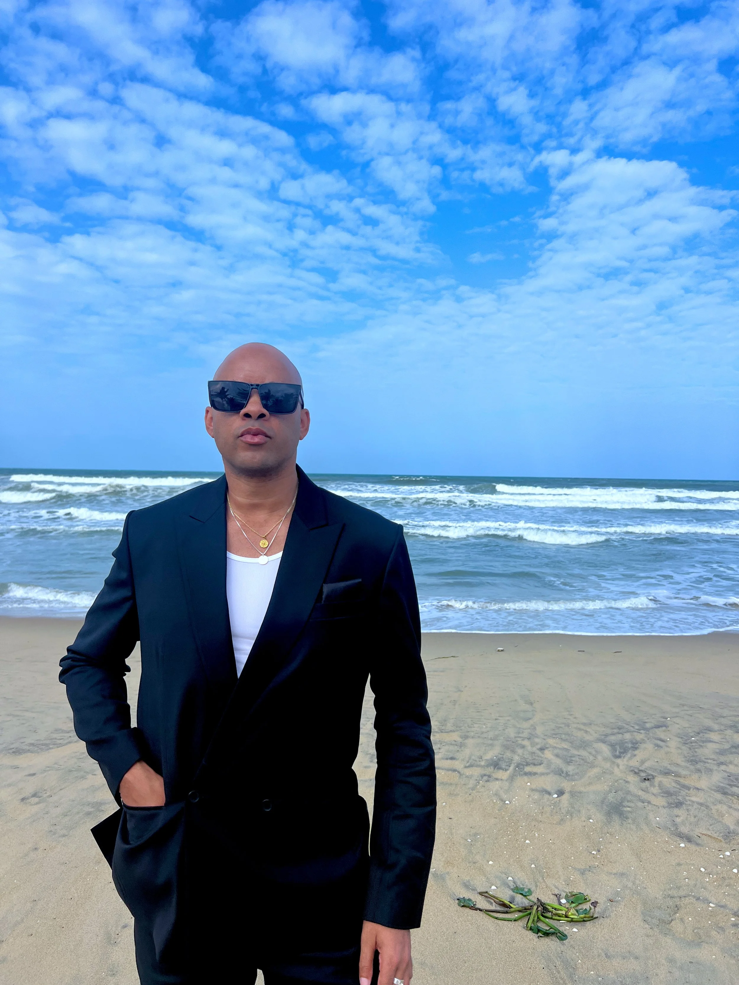 Ethan Ward stands facing the camera on a beach, dressed in a black blazer, white shirt, and sunglasses, with rolling ocean waves and a vivid blue, cloud-filled sky in the background.