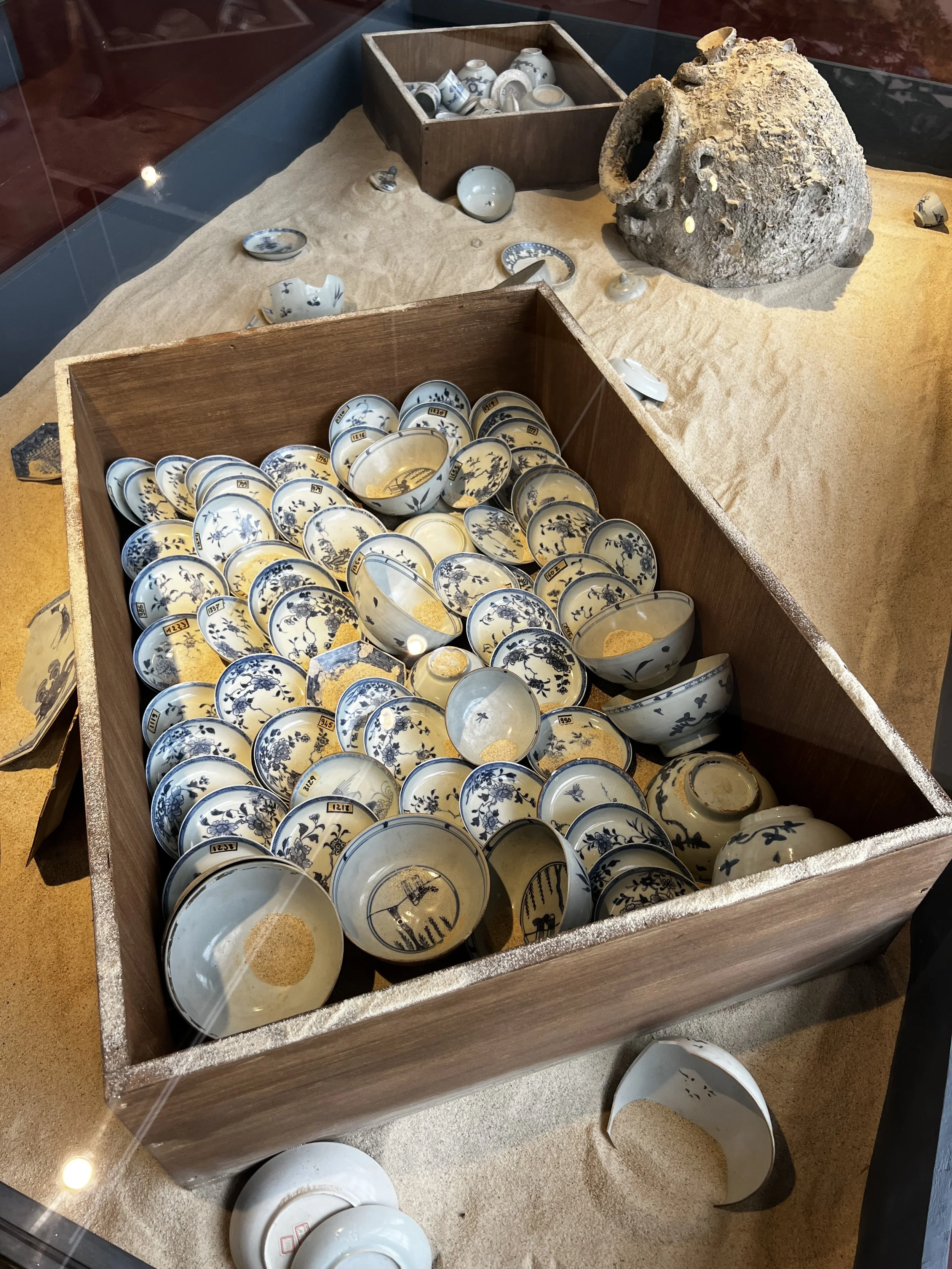 A wooden display box filled with broken blue and white porcelain bowls with intricate designs, some scattered on a yellowish sand-like surface, with a large weathered ceramic vessel and smaller porcelain pieces nearby.