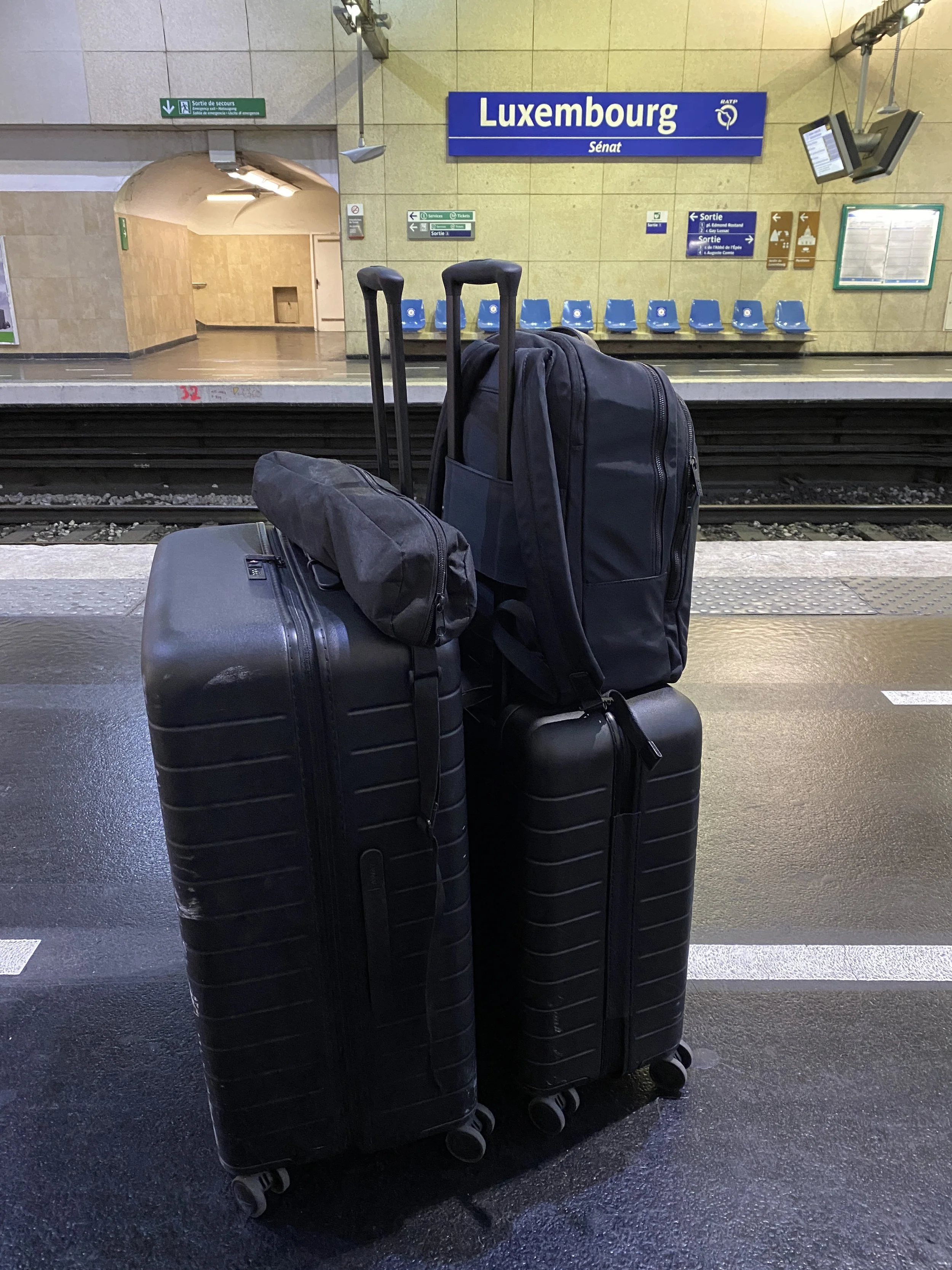 Travel luggage consisting of three black suitcases and a black backpack standing on a train station platform with a Luxembourg sign and empty blue seats in the background.