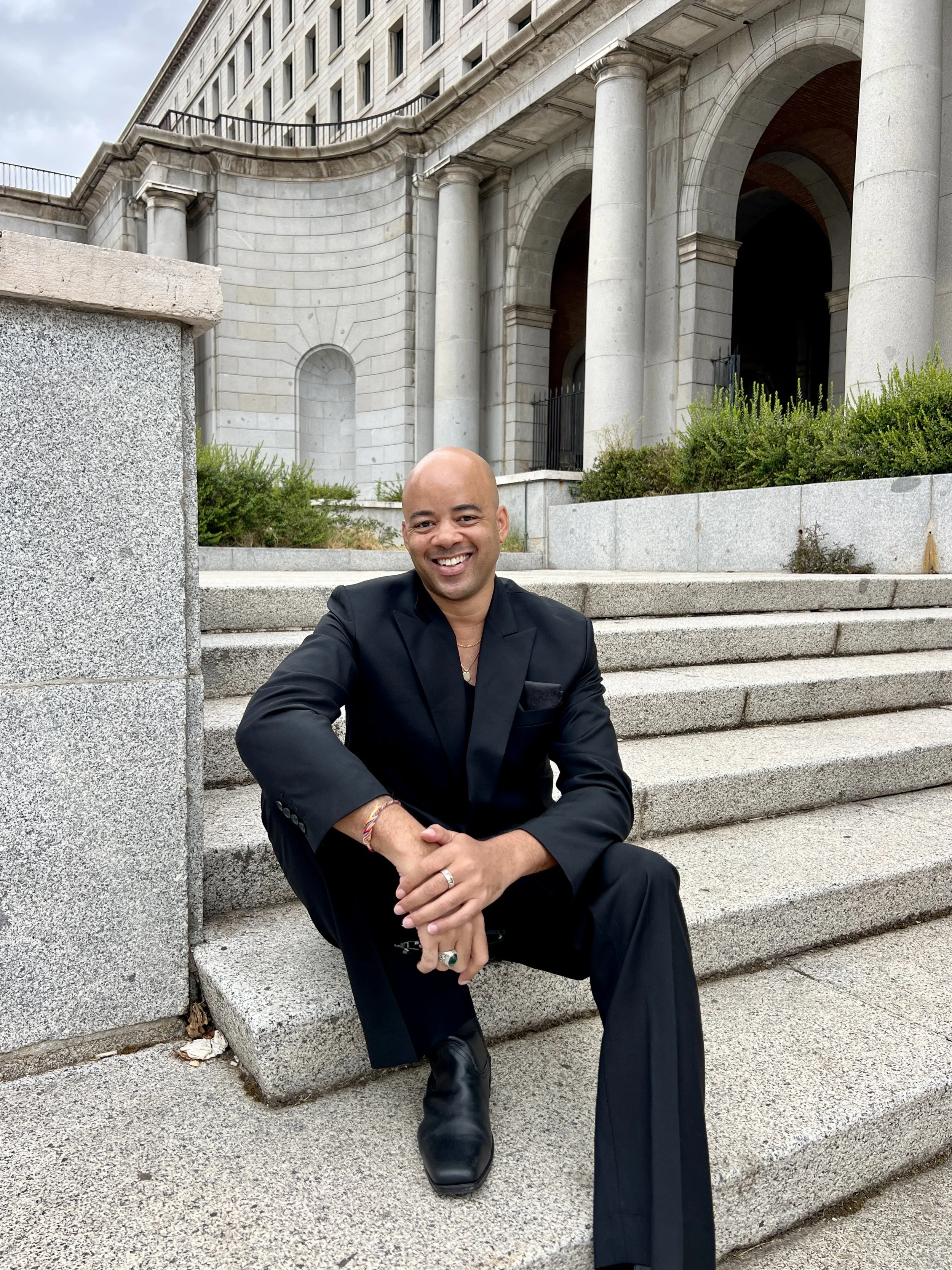 Ethan Ward sits on granite steps wearing an all black suit and smiling at the camera.