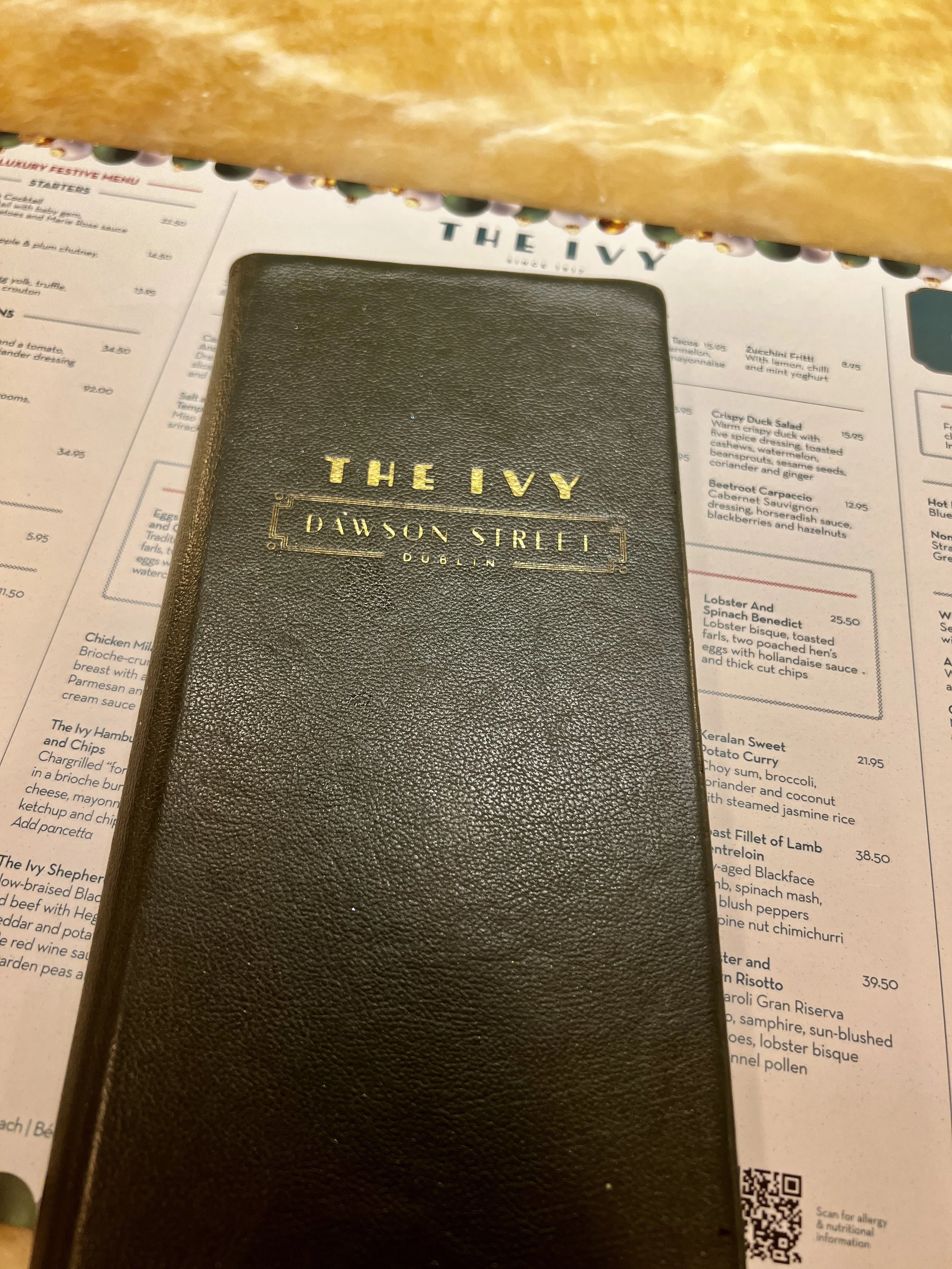 Black leather menu cover with gold lettering reading "The Ivy Dawson Street Dublin" placed on a restaurant menu, with a bread basket partially visible at the top.