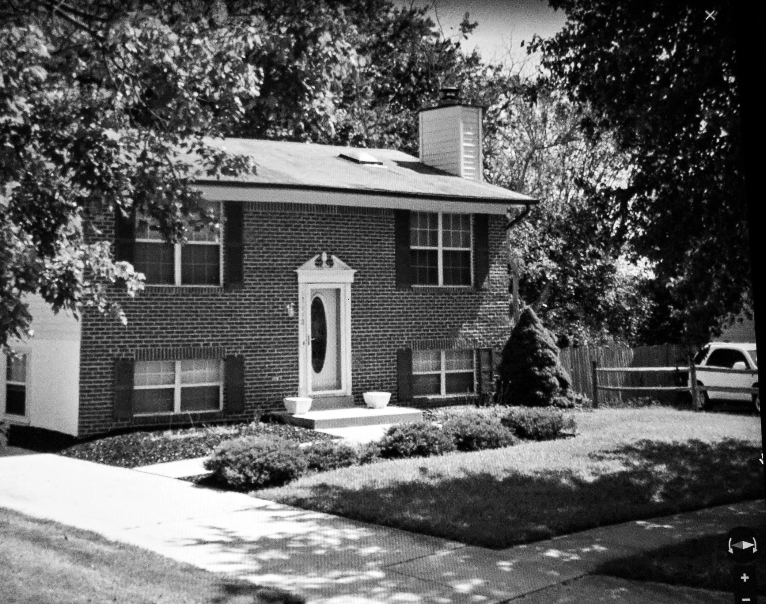 A two-story brick house with a front porch and a white door, surrounded by trees and a well-kept lawn, with a sidewalk and parked cars visible.