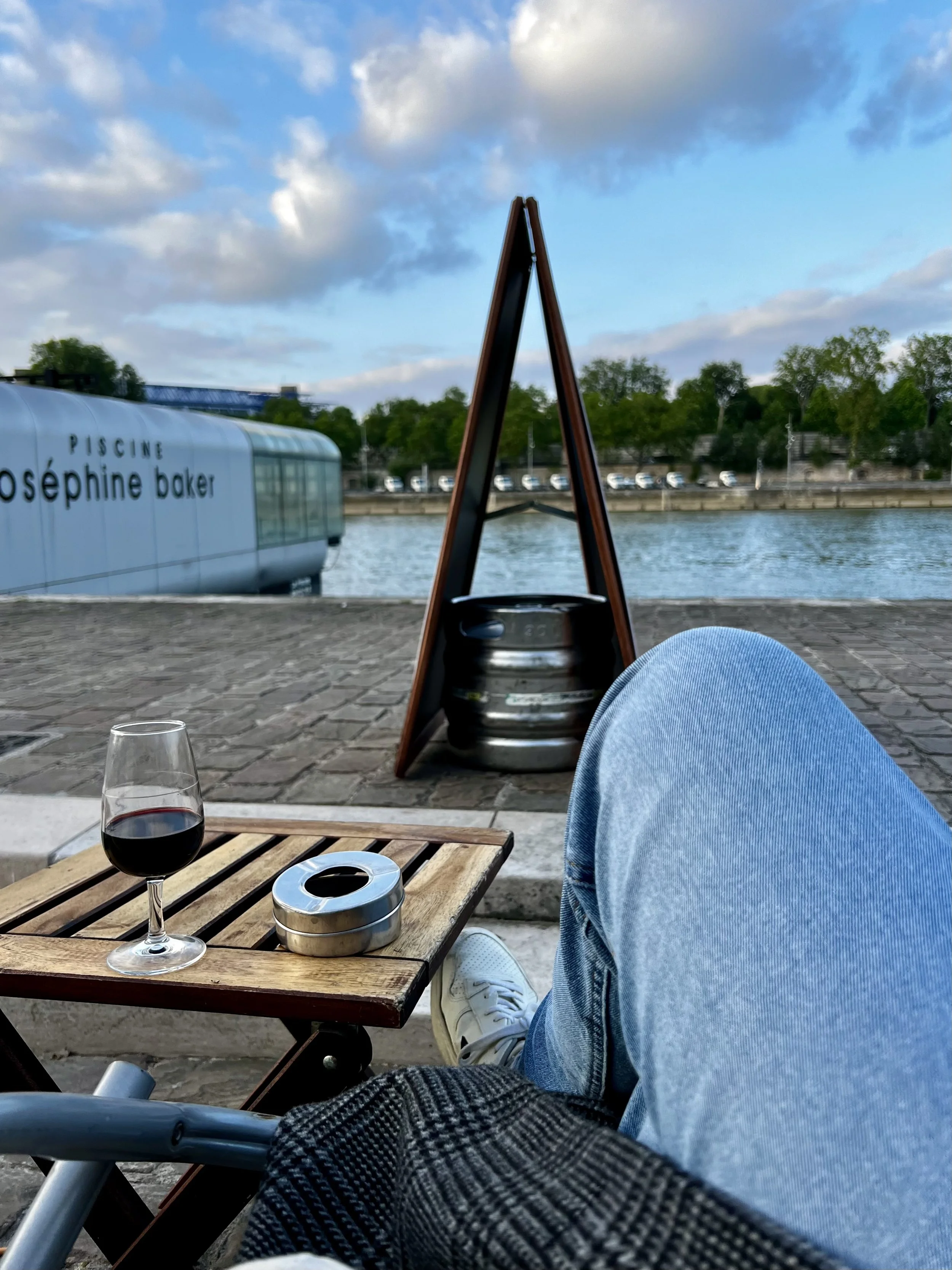 Person sitting near a river with a glass of red wine, a small wooden table, and a beer keg with a chalkboard sign. In the background, there's a modern building labeled 'Piscine Joséphine Baker' and trees along the riverbank, under a partly cloudy sky