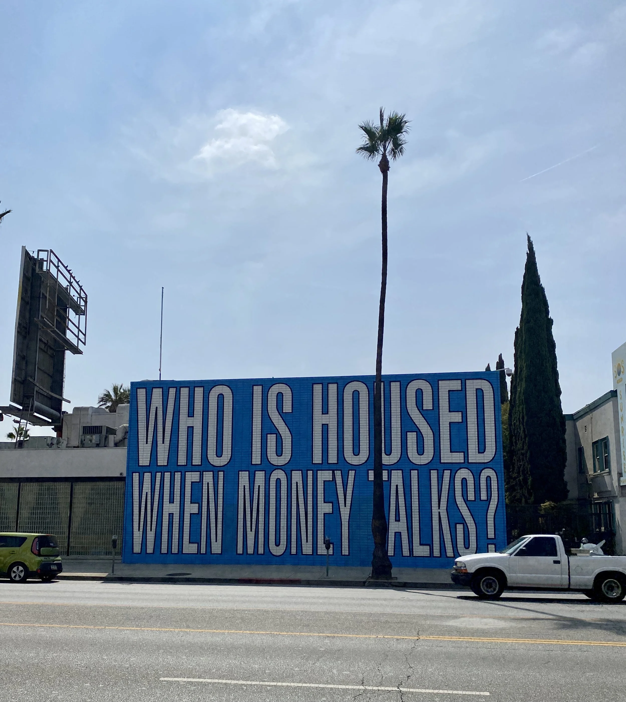A large banner with the text 'WHO IS HOUSED WHEN MONEY TALKS?' displayed on a building in an urban area with cars parked in front, palm trees, and a partly cloudy sky.