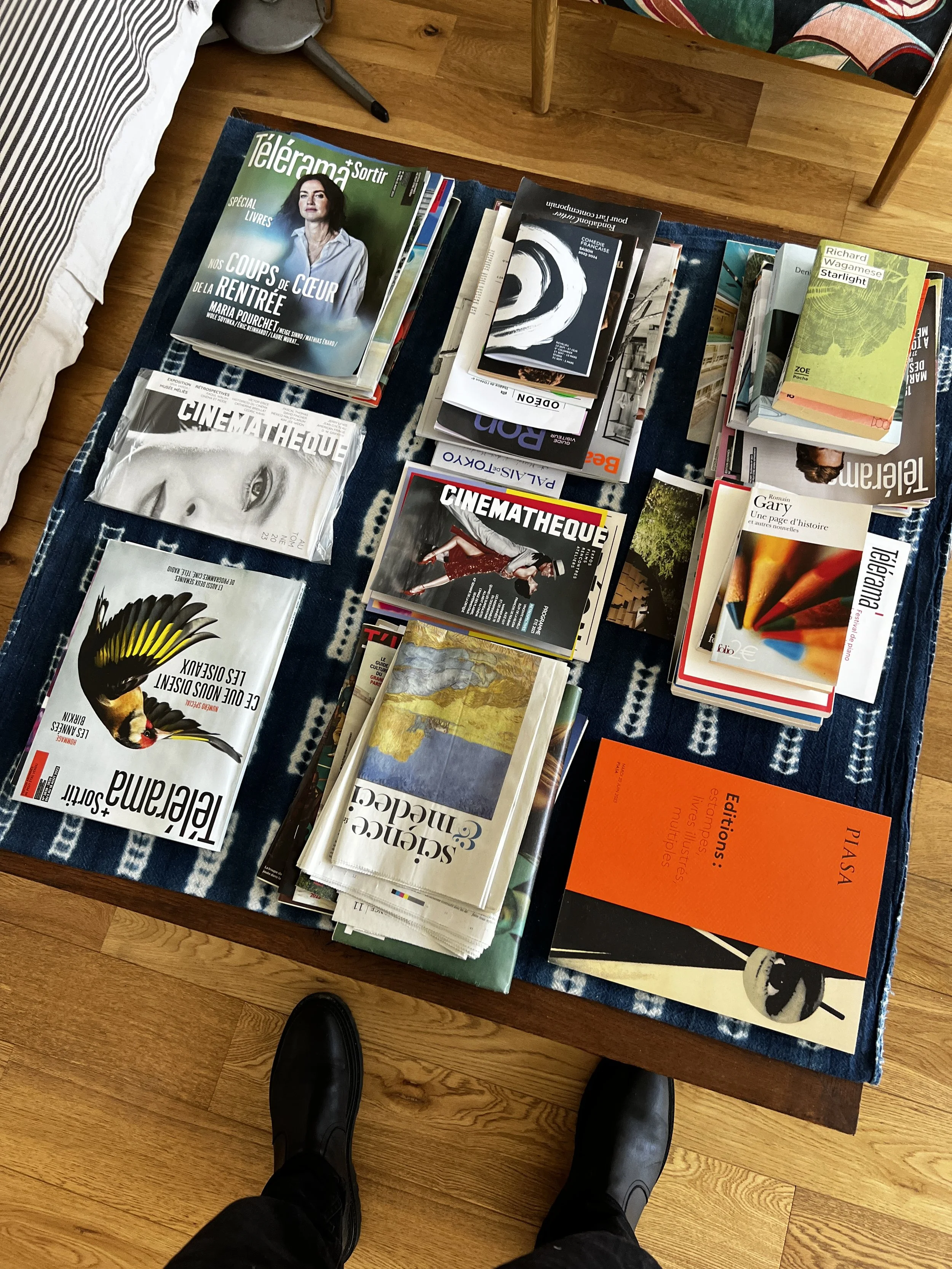 A top-down view of a wooden table covered with stacks of magazines and books on art, cinema, poetry, and culture in a room with hardwood flooring. The photographer's shoes are visible at the bottom of the image.