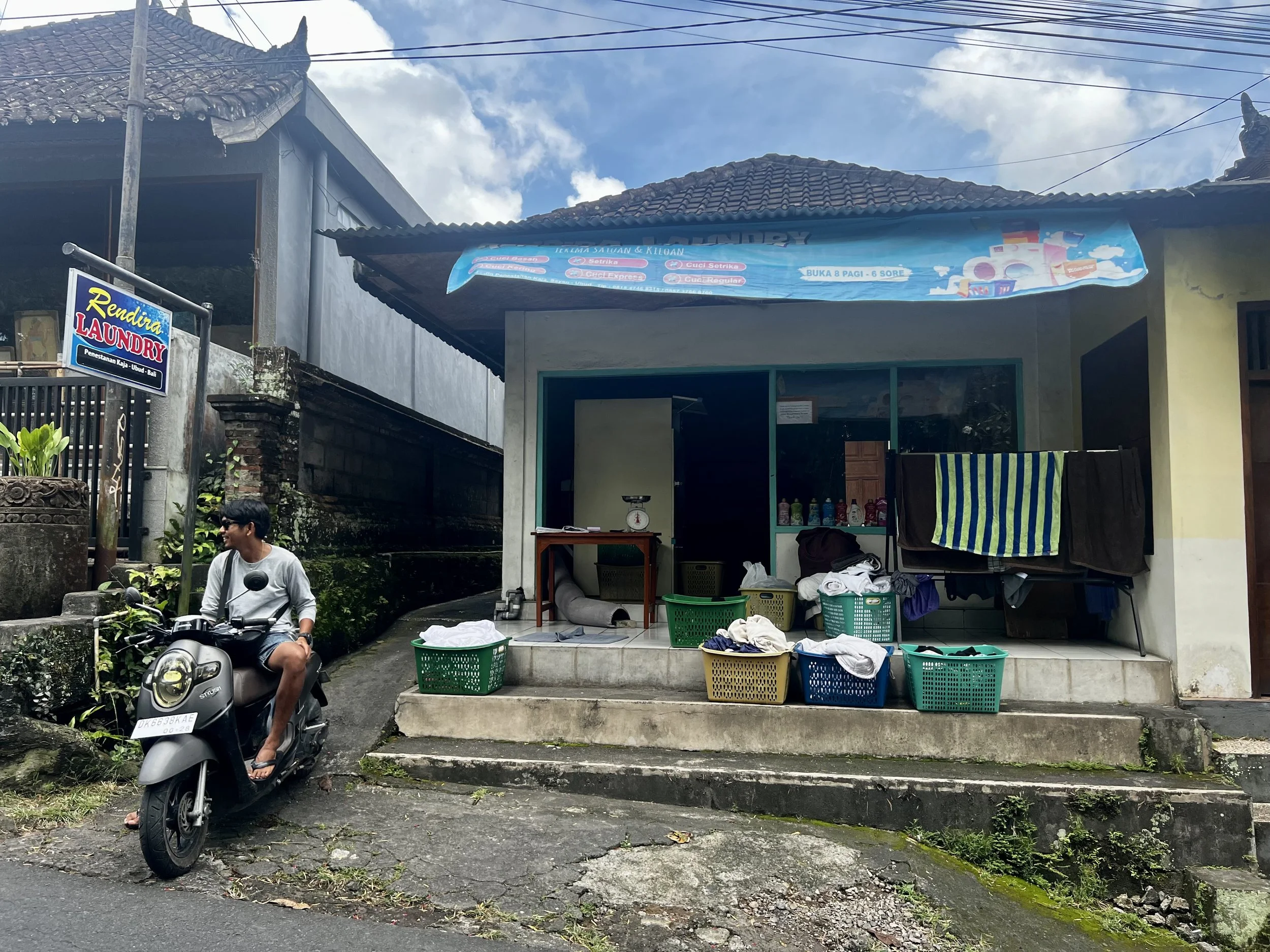 A small laundry shop with laundry baskets filled with clothes, a drying rack with clothes, and a man sitting on a motorcycle outside.