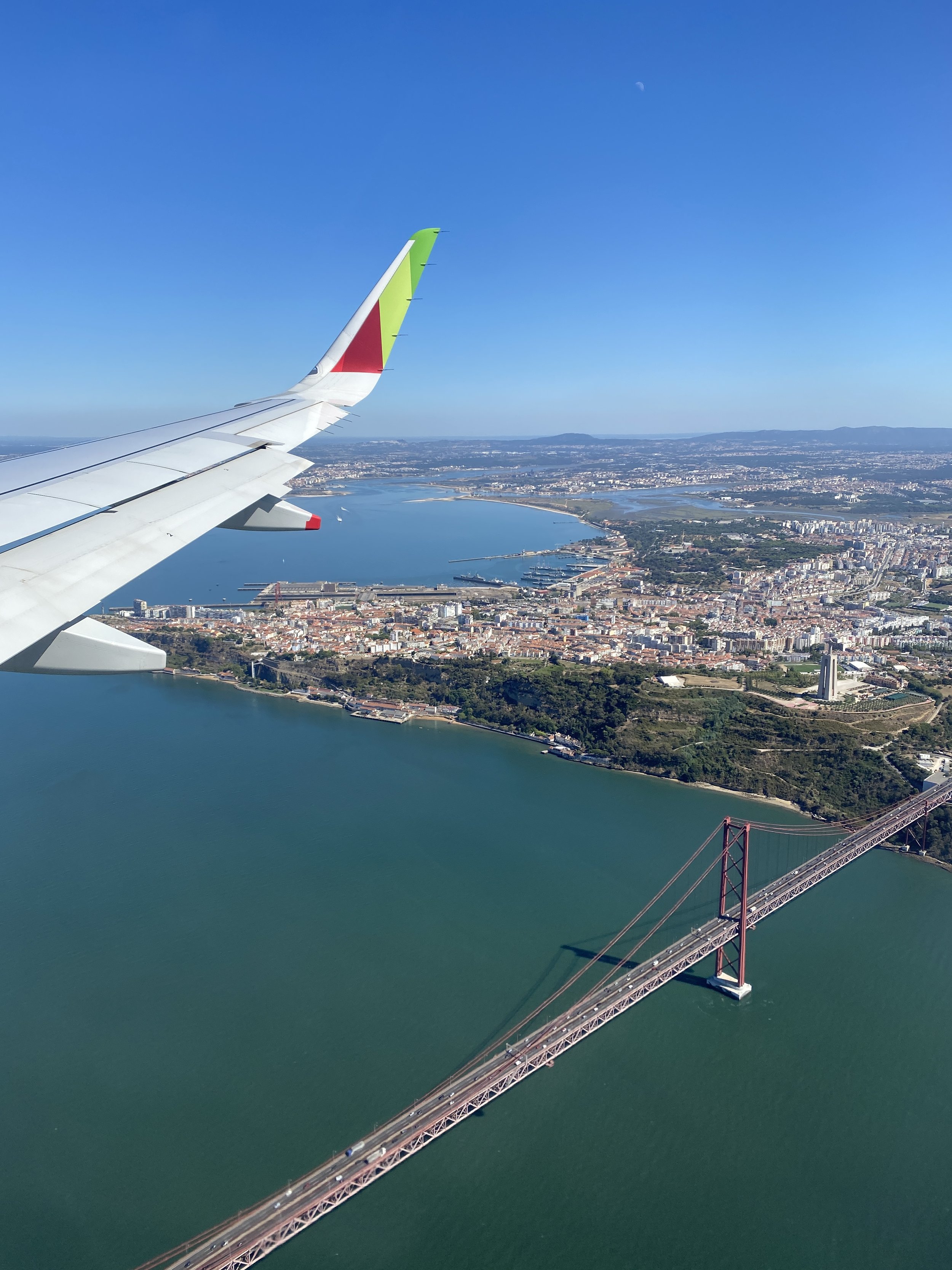 View from an airplane window showing a city with a large bay, a bridge over green water, and an airplane wing with a colorful logo.