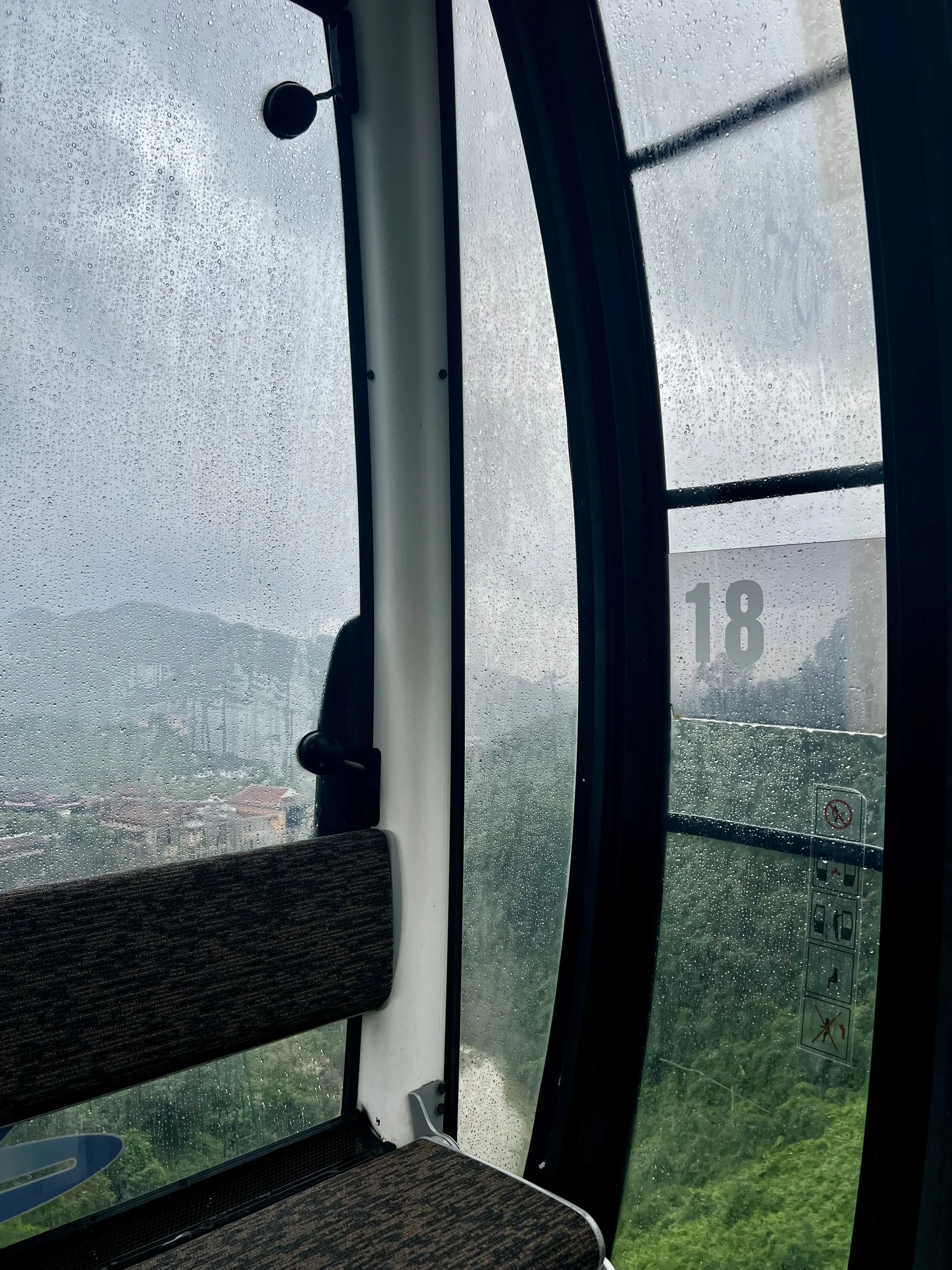 View from inside a ferris wheel cabin with rain-soaked glass, showing a cloudy sky, green landscape, and distant mountains.