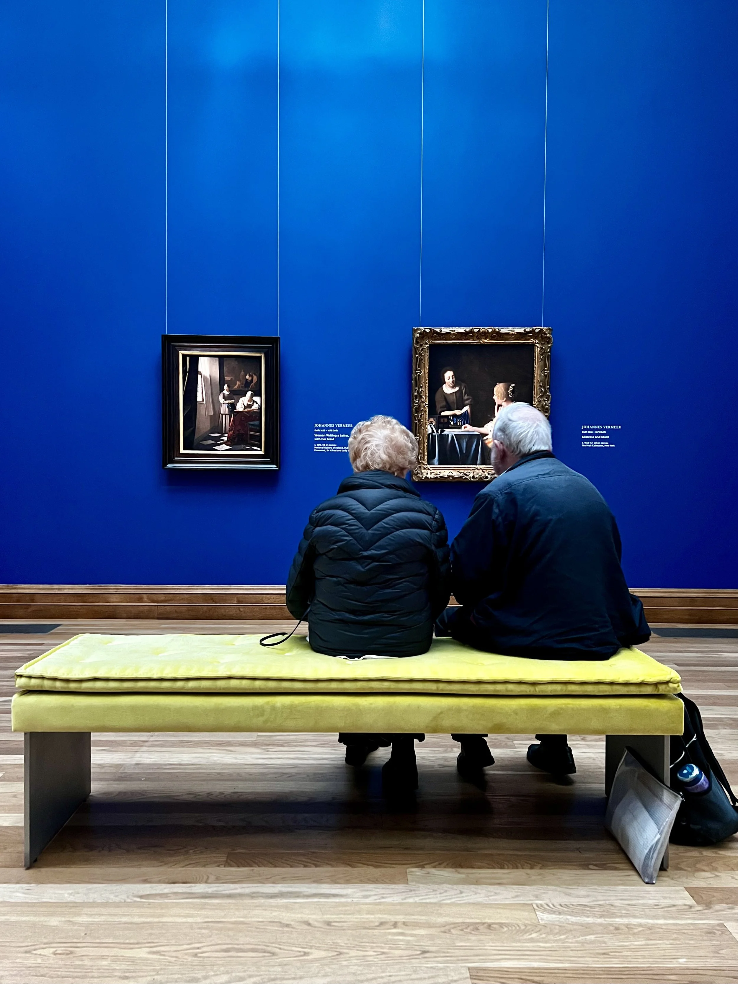 Two elderly people sitting on a yellow bench, observing paintings at an art gallery with a vibrant blue wall behind them.