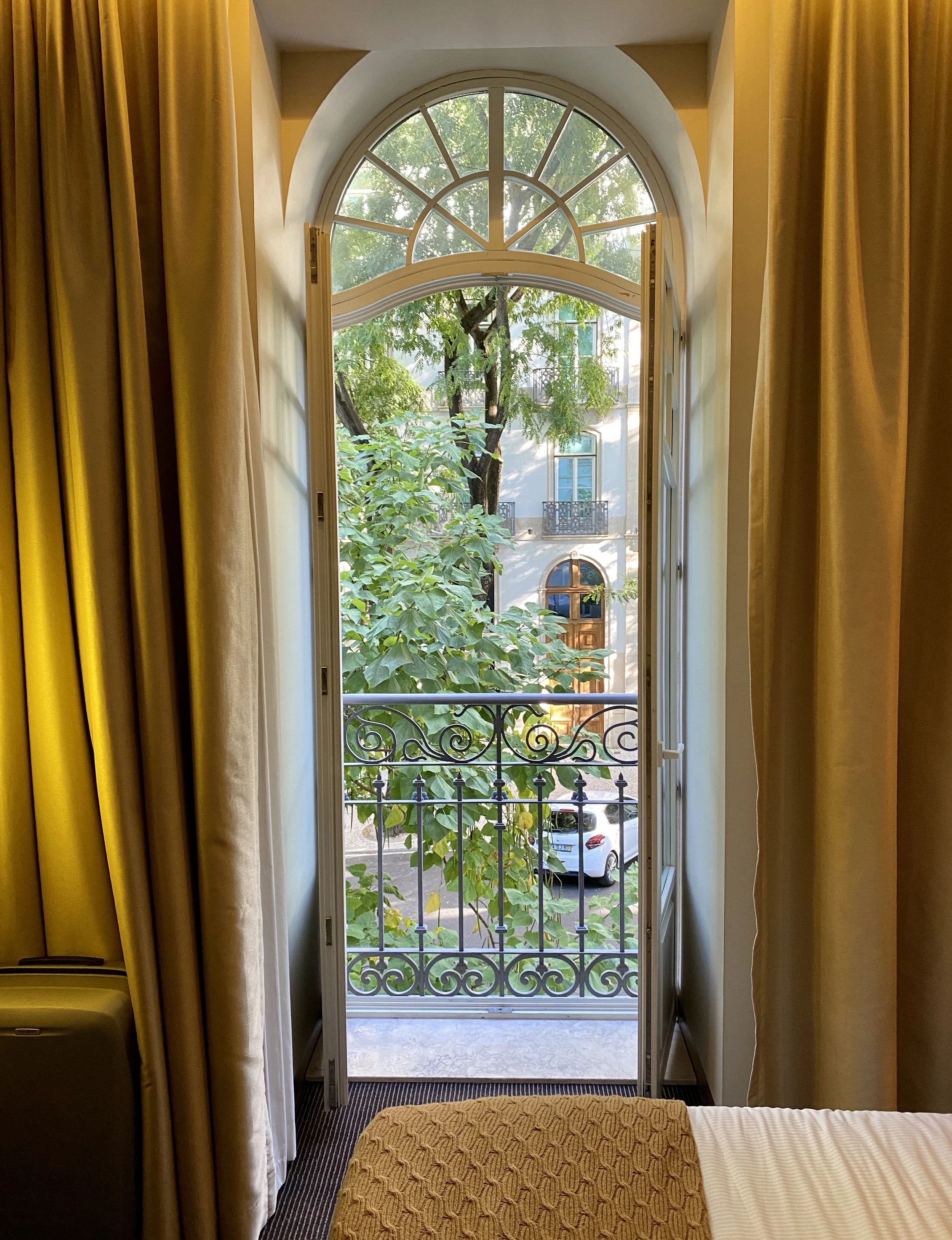Open French door leading to small balcony with decorative iron railing, outside view of green trees, neighboring building with arched window and balcony, parked cars, room with yellow curtains and part of bed with textured bedspread.