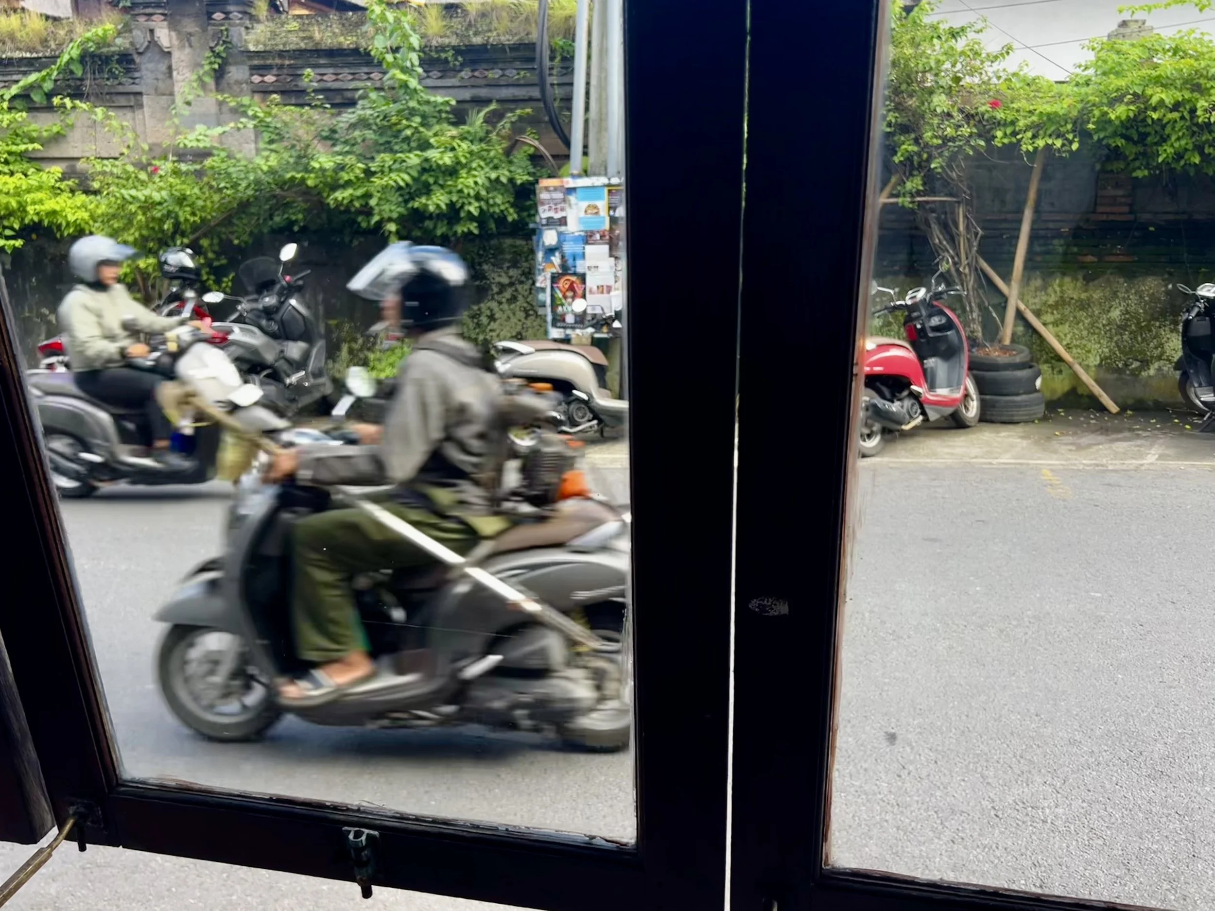 View through glass door showing motorbikes and scooters parked outside on the street with two people riding scooters passing by.