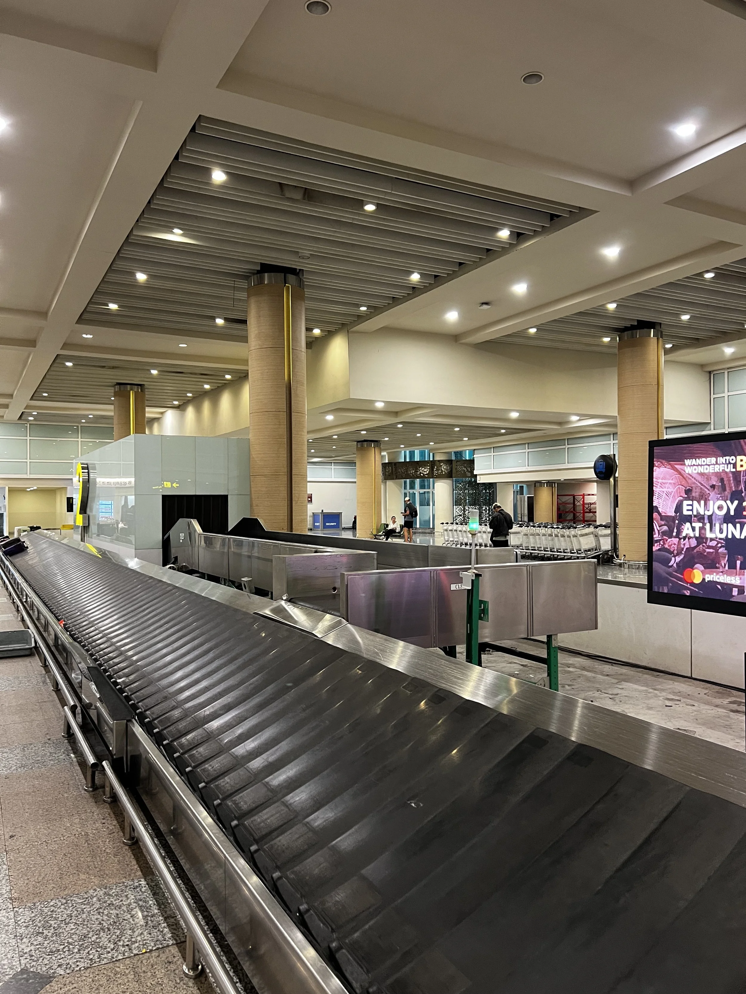 Empty baggage claim area at an airport with a curved conveyor belt, illuminated ceiling, and a digital advertising screen.