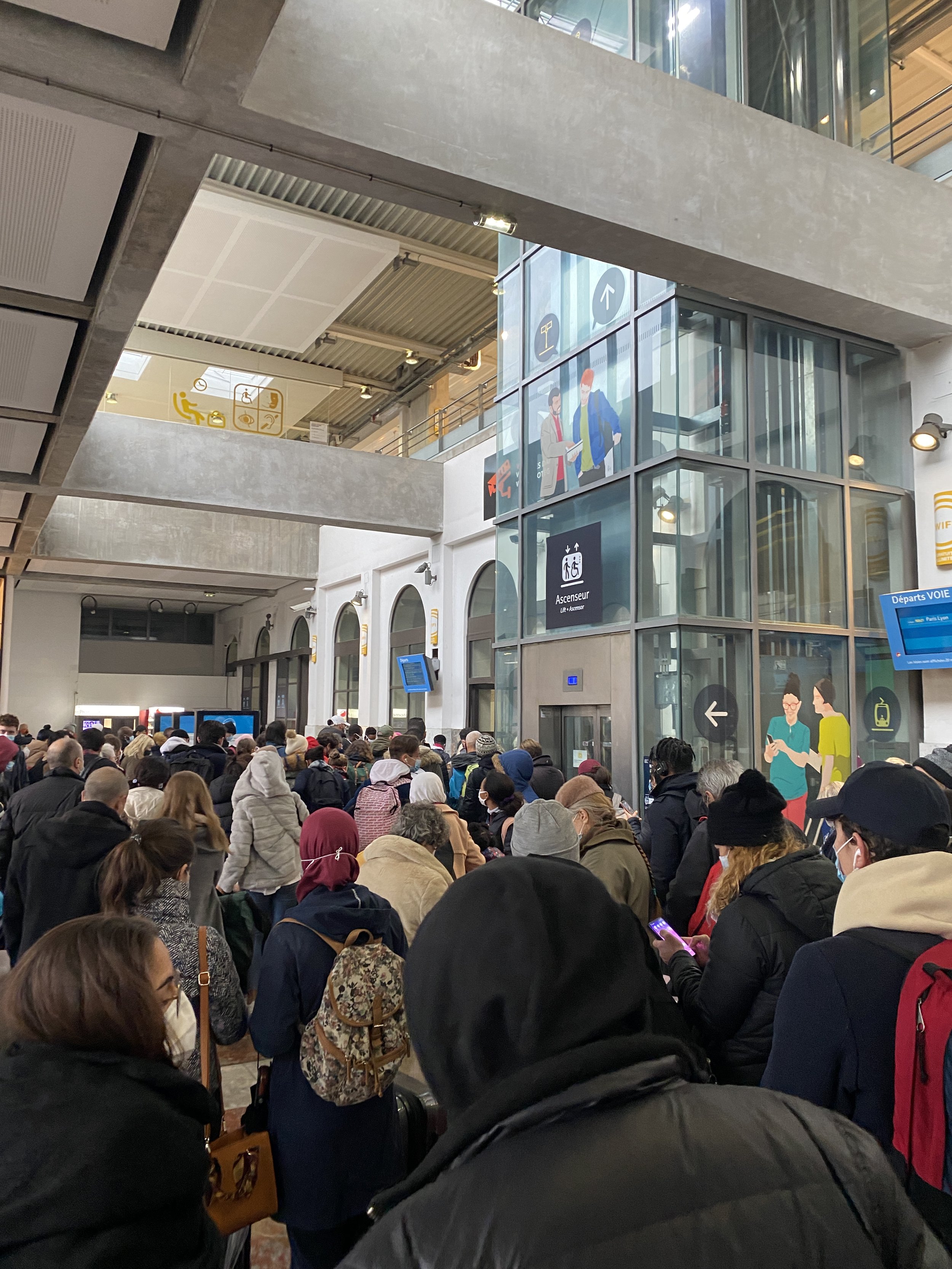 A busy scene inside a train station with a large crowd of people waiting. The station has modern architecture with concrete and glass elements, digital screens, and signs indicating directions and services.