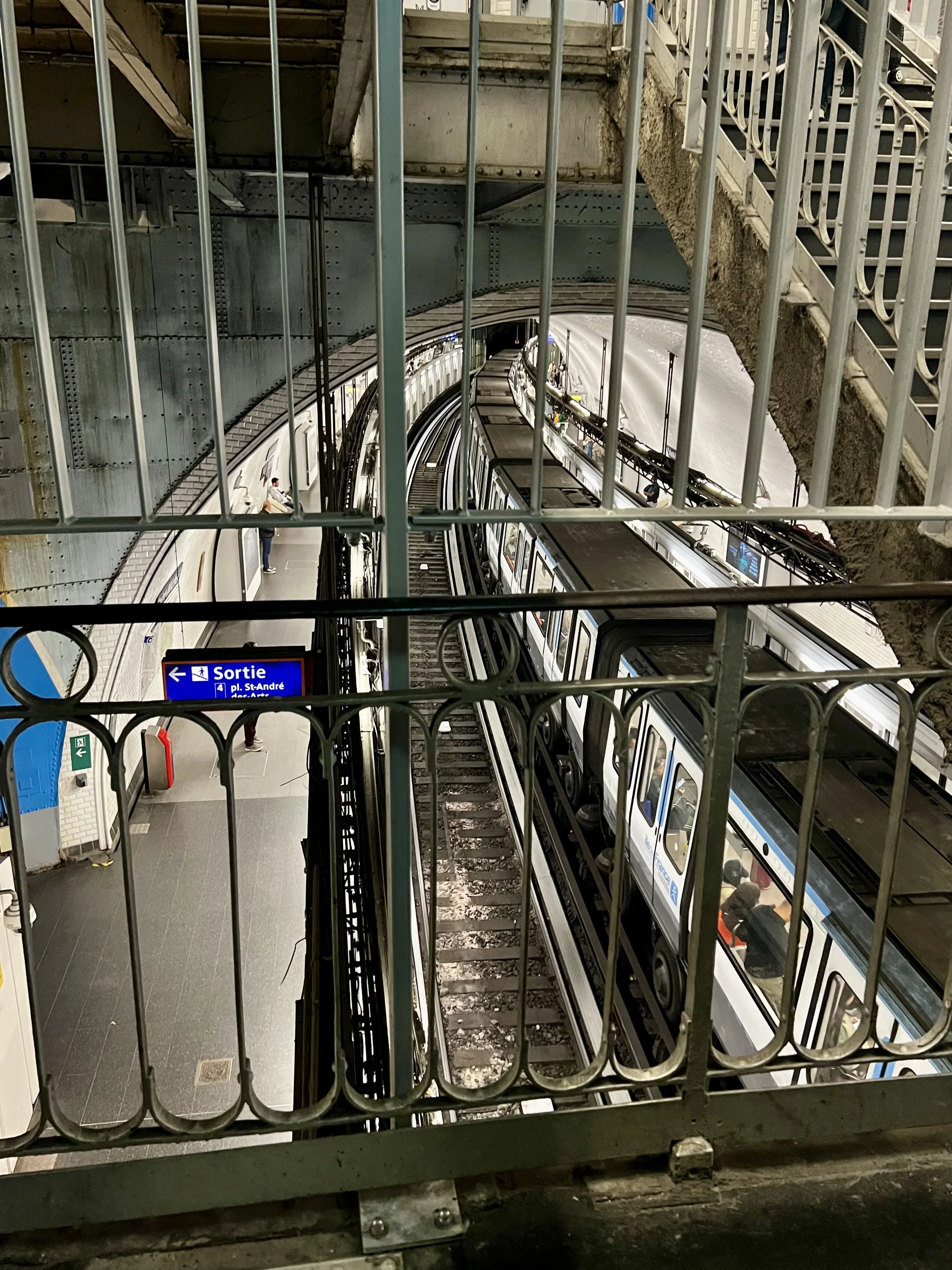 View of an underground subway station platform with a subway train on the tracks, seen through a metal railing. There is a sign with the word 'Sortie' indicating an exit, and a few passengers are waiting or walking on the platform.