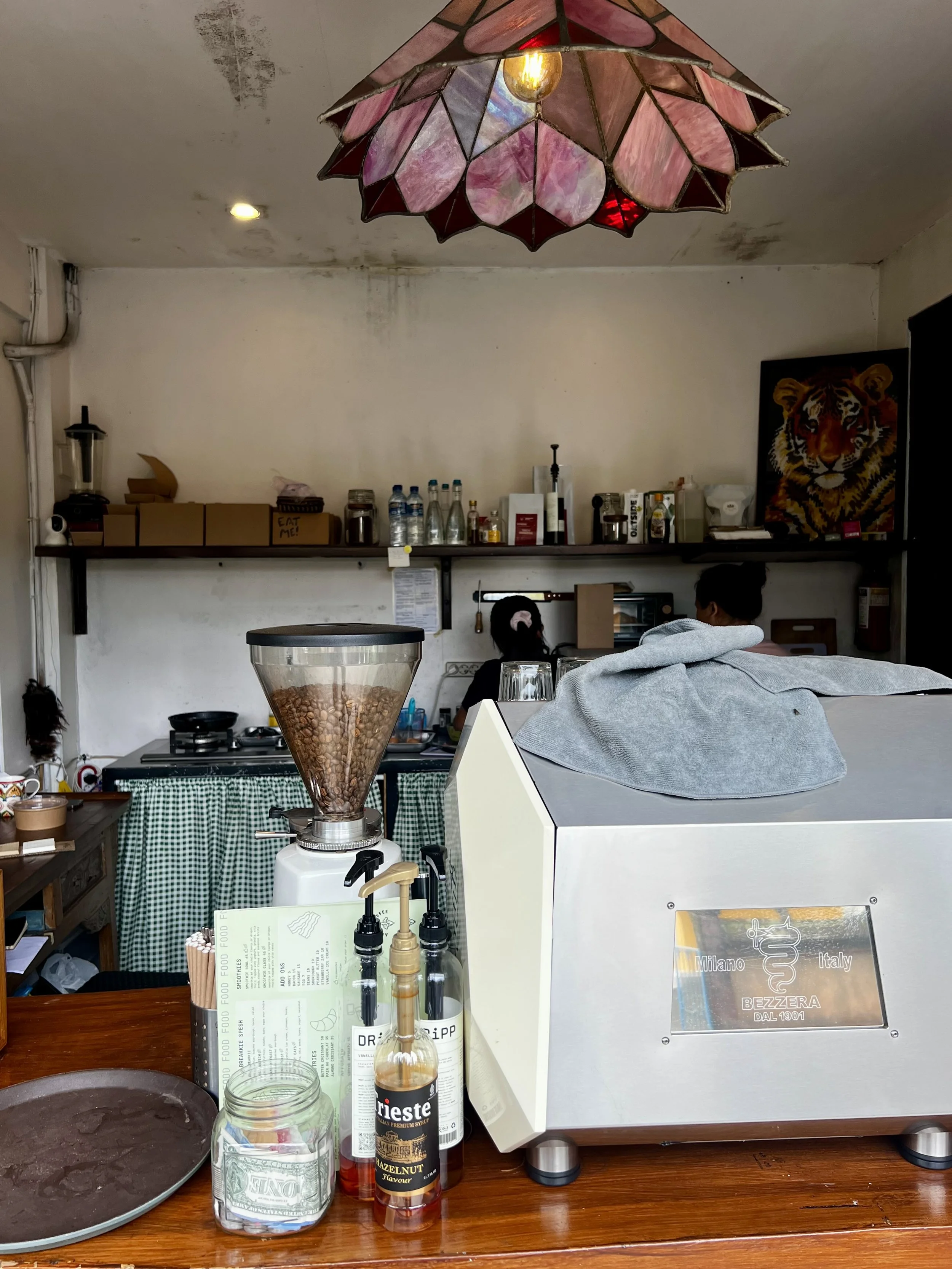 Interior of a coffee shop with a coffee grinder, bottles, and coffee supplies on the counter, two people working in the background, and a decorative pink stained glass ceiling lamp.