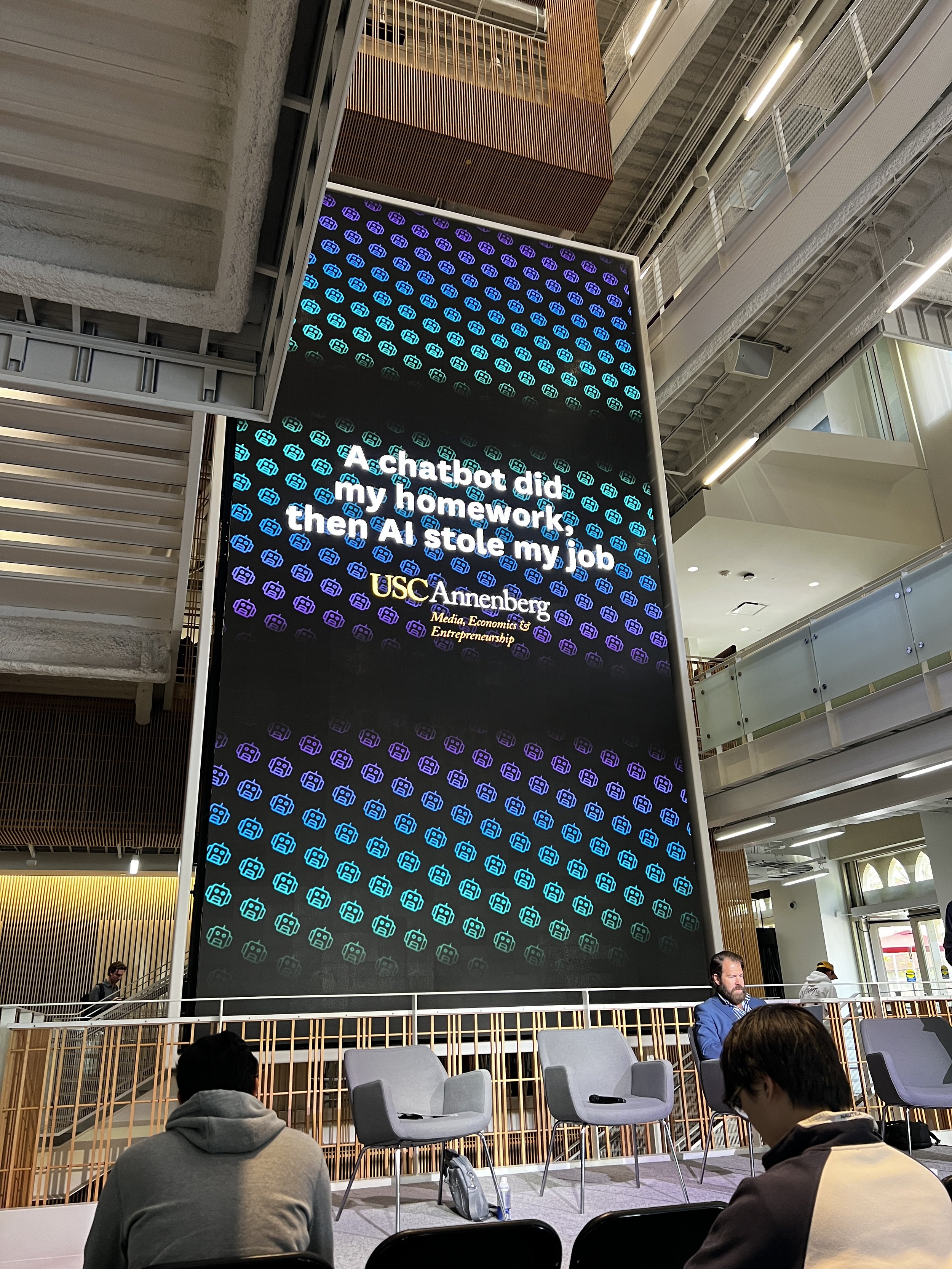 Large digital screen at USC Annenberg School displaying a quote about a chatbot and AI, with chairs and people sitting in front of it inside a modern building.