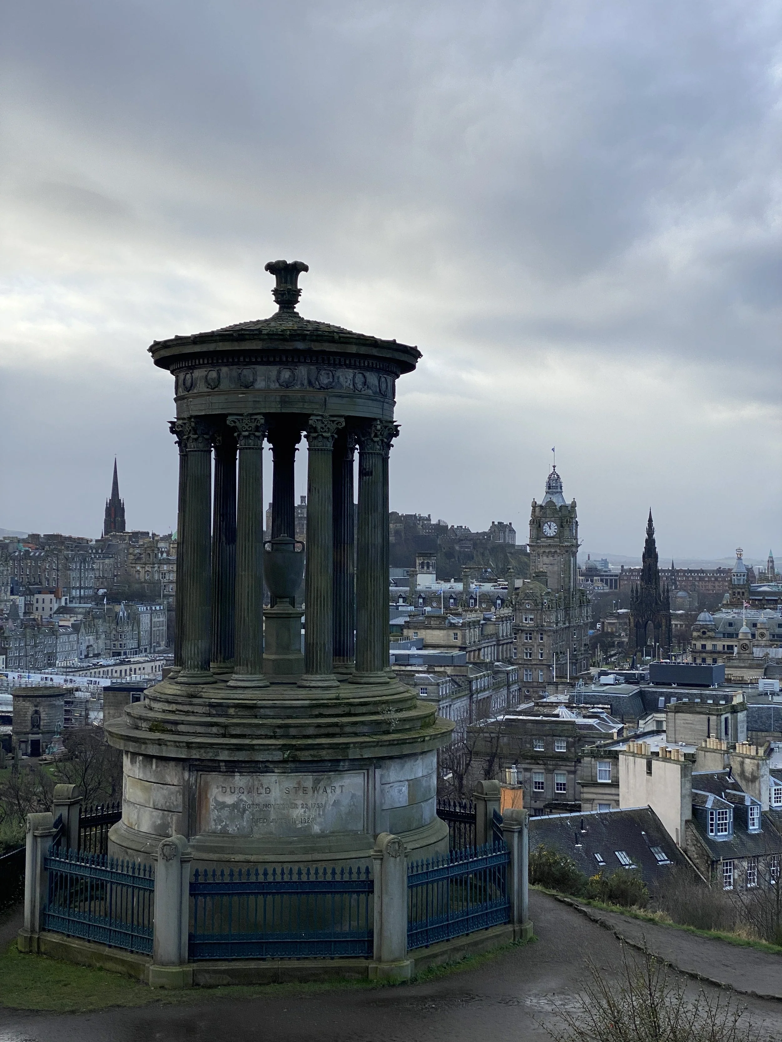 Historical monument with columns near a cityscape, featuring clock towers and numerous buildings during overcast weather.