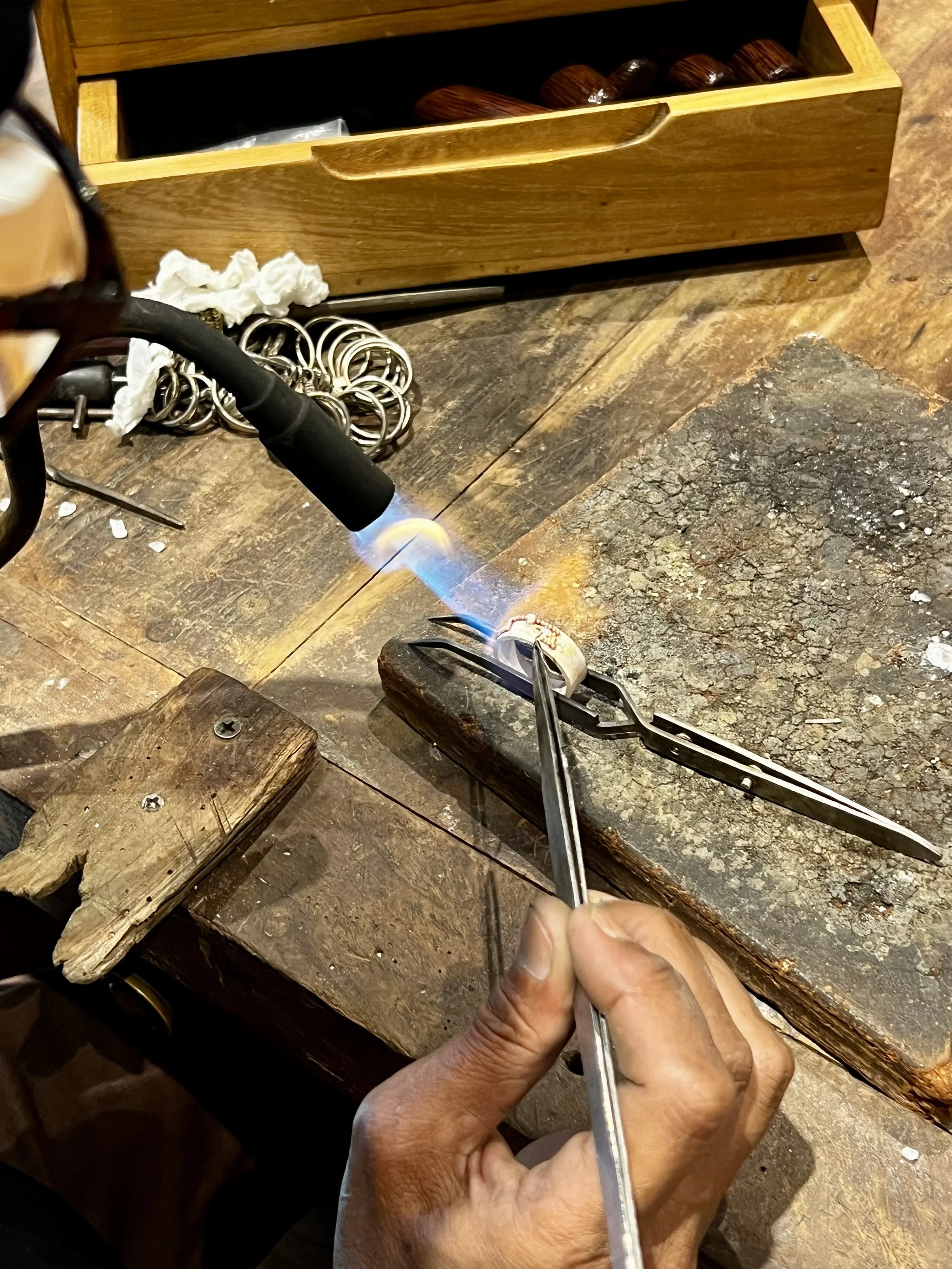 A person working with a blowtorch on a metal piece in a workshop, with tools, jewelry rings, and boxes nearby.