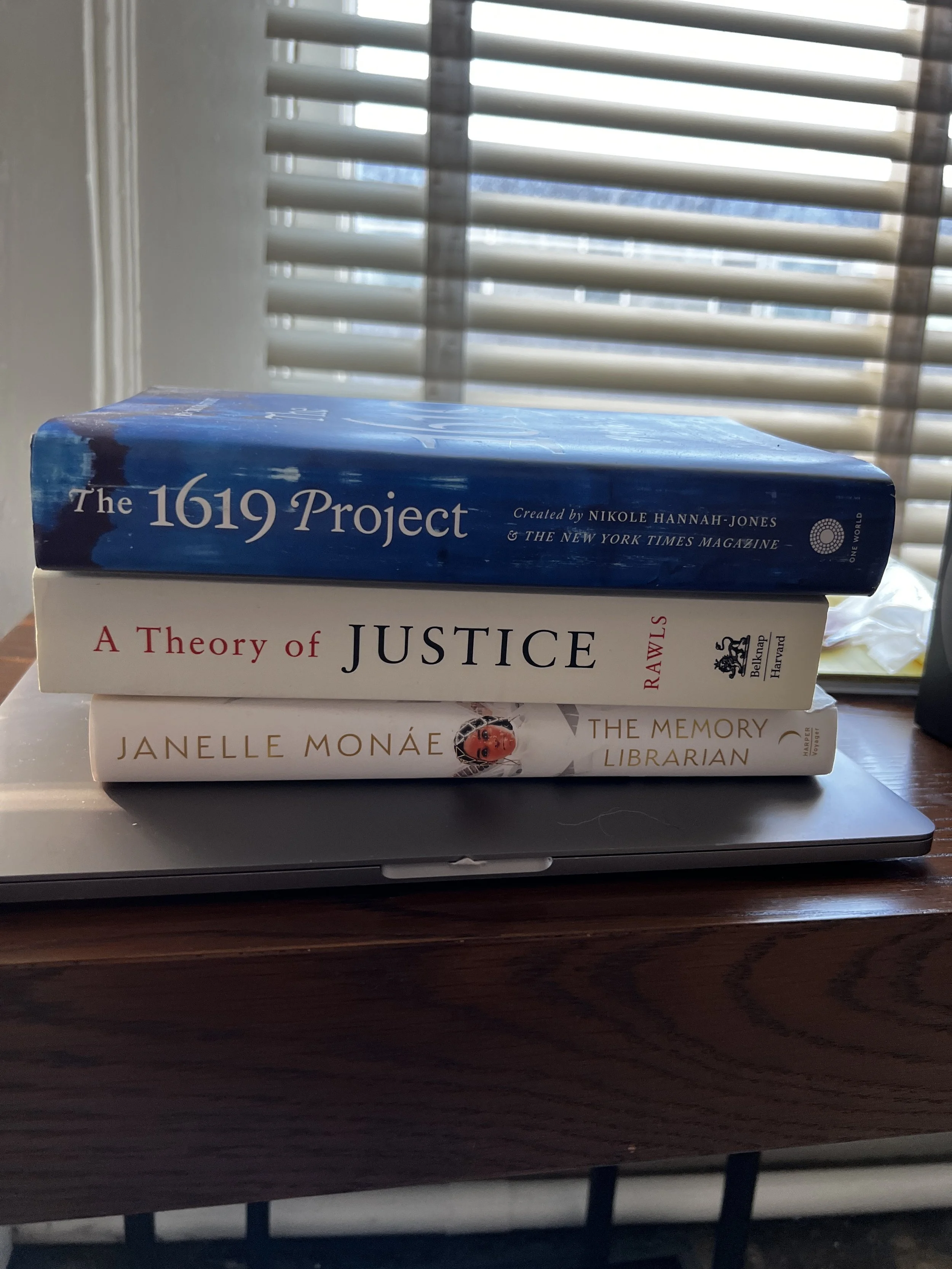 A stack of three books on a laptop, with a window and blinds in the background. The top book is titled 'The 1619 Project,' the middle book is 'A Theory of Justice,' and the bottom book is 'The Memory Librarian' by Janelle Monáe.