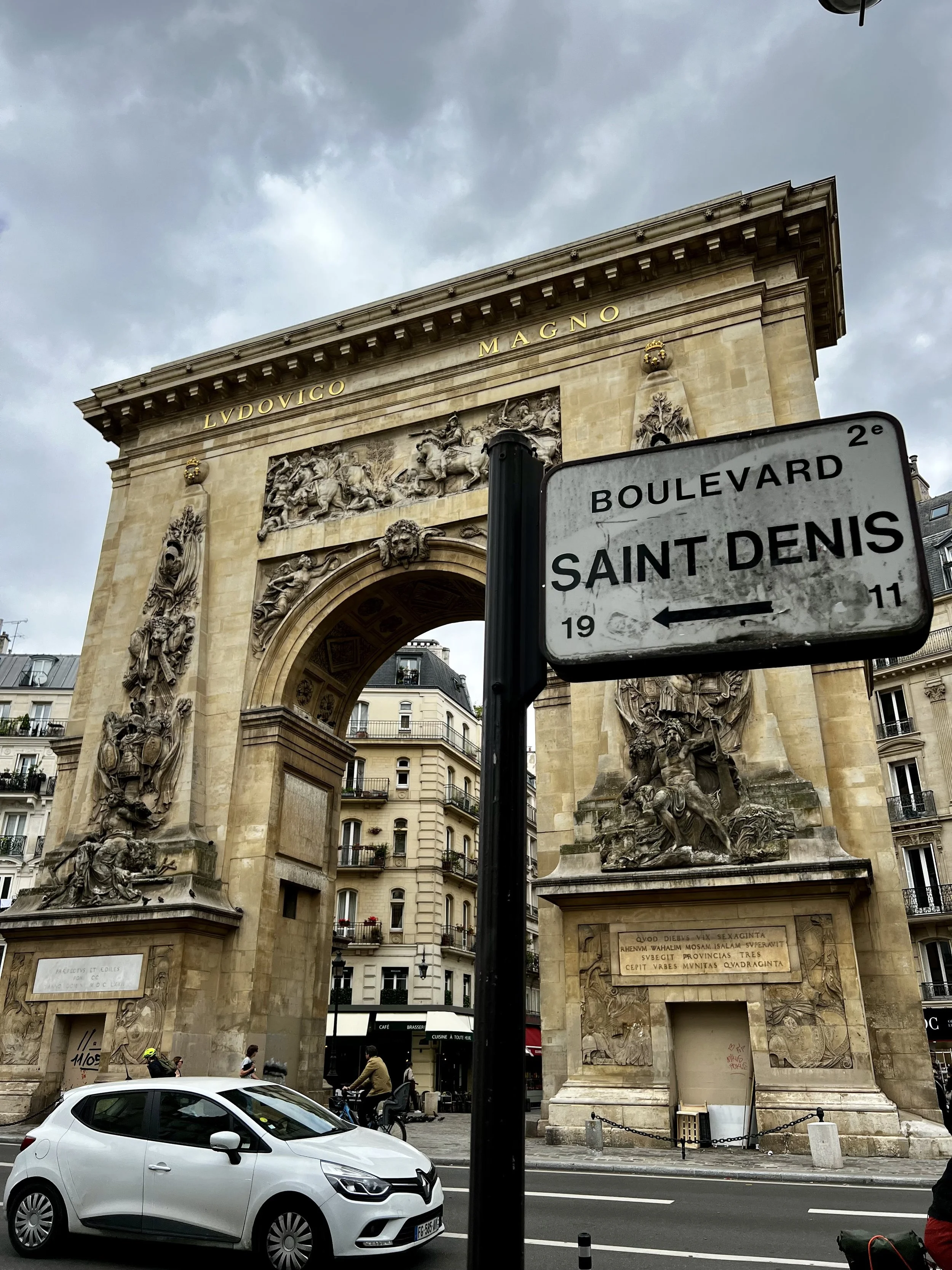 A historic archway with intricate sculptures and gold lettering up top, framed by a street scene with a white car and pedestrians. A street sign points to Saint Denis boulevard.