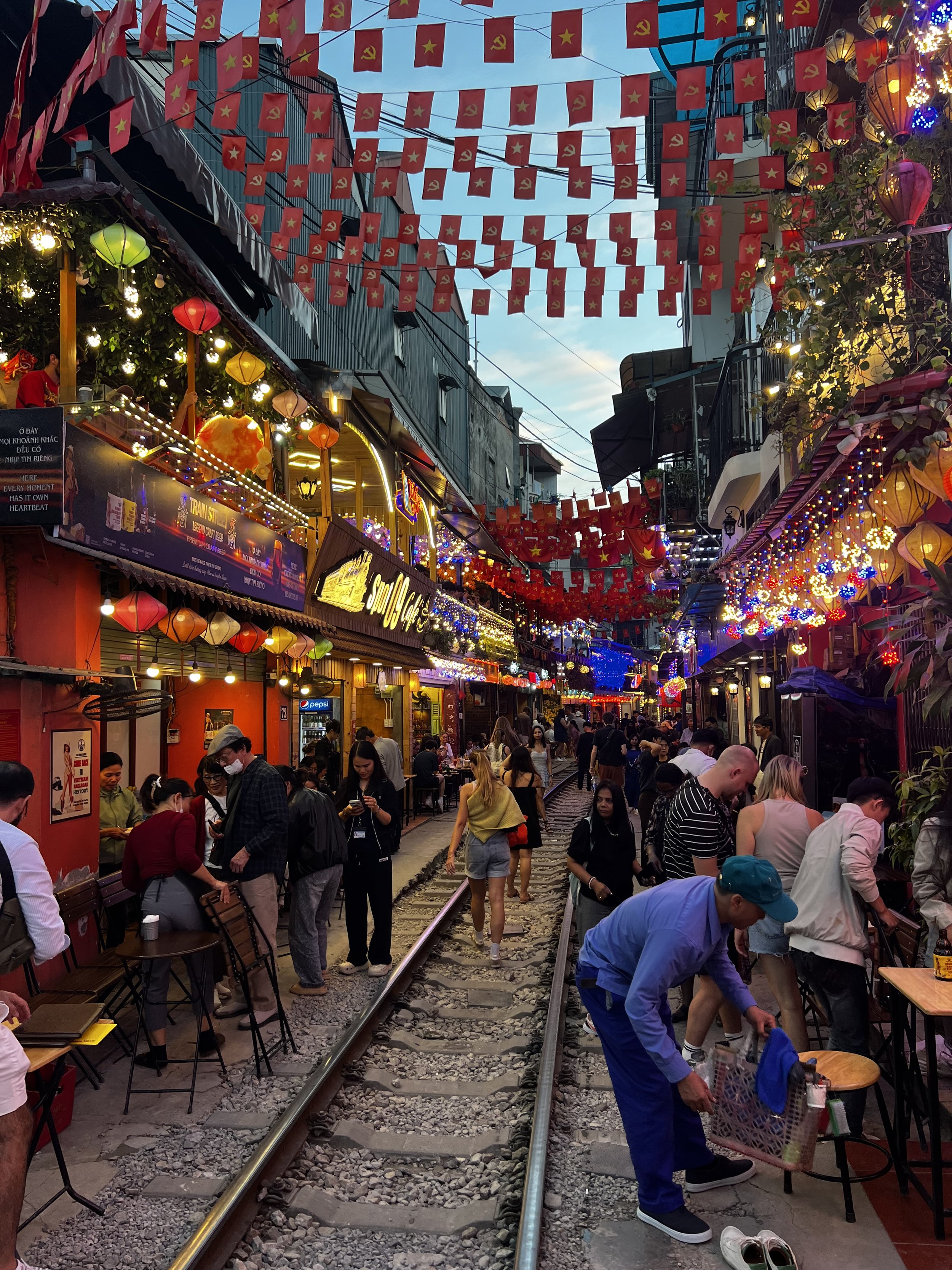 Crowded street with shops and restaurants along both sides of a railway track, decorated with colorful lanterns and flags, with people walking and sitting at outdoor tables during evening.