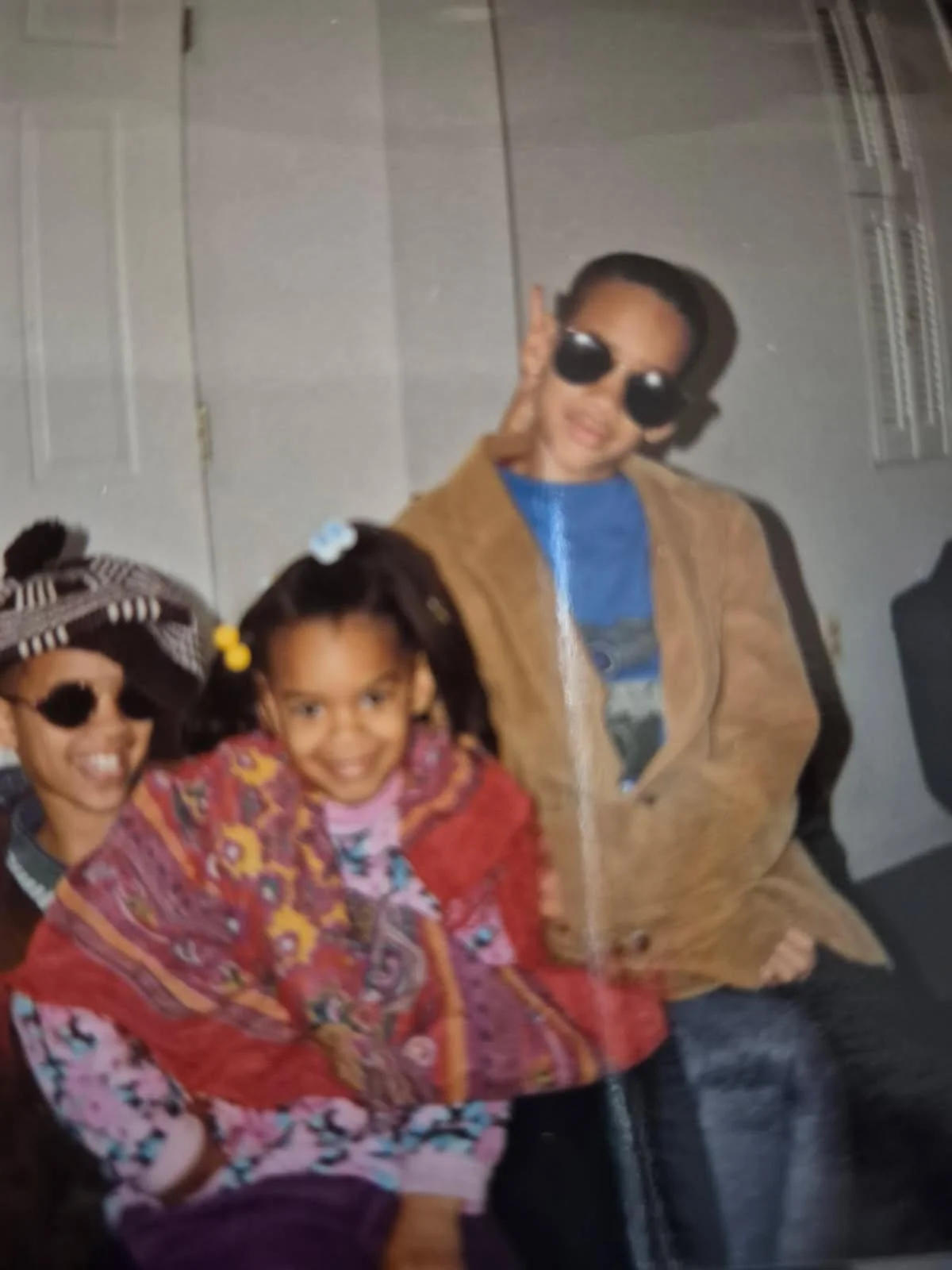 sitting side by side, smiling, wearing sunglasses, with a gray wall in the background. One person is a young girl with pigtails wearing a colorful jacket, the second is a woman with a headscarf and glasses, and the third is a man wearing a brown coat