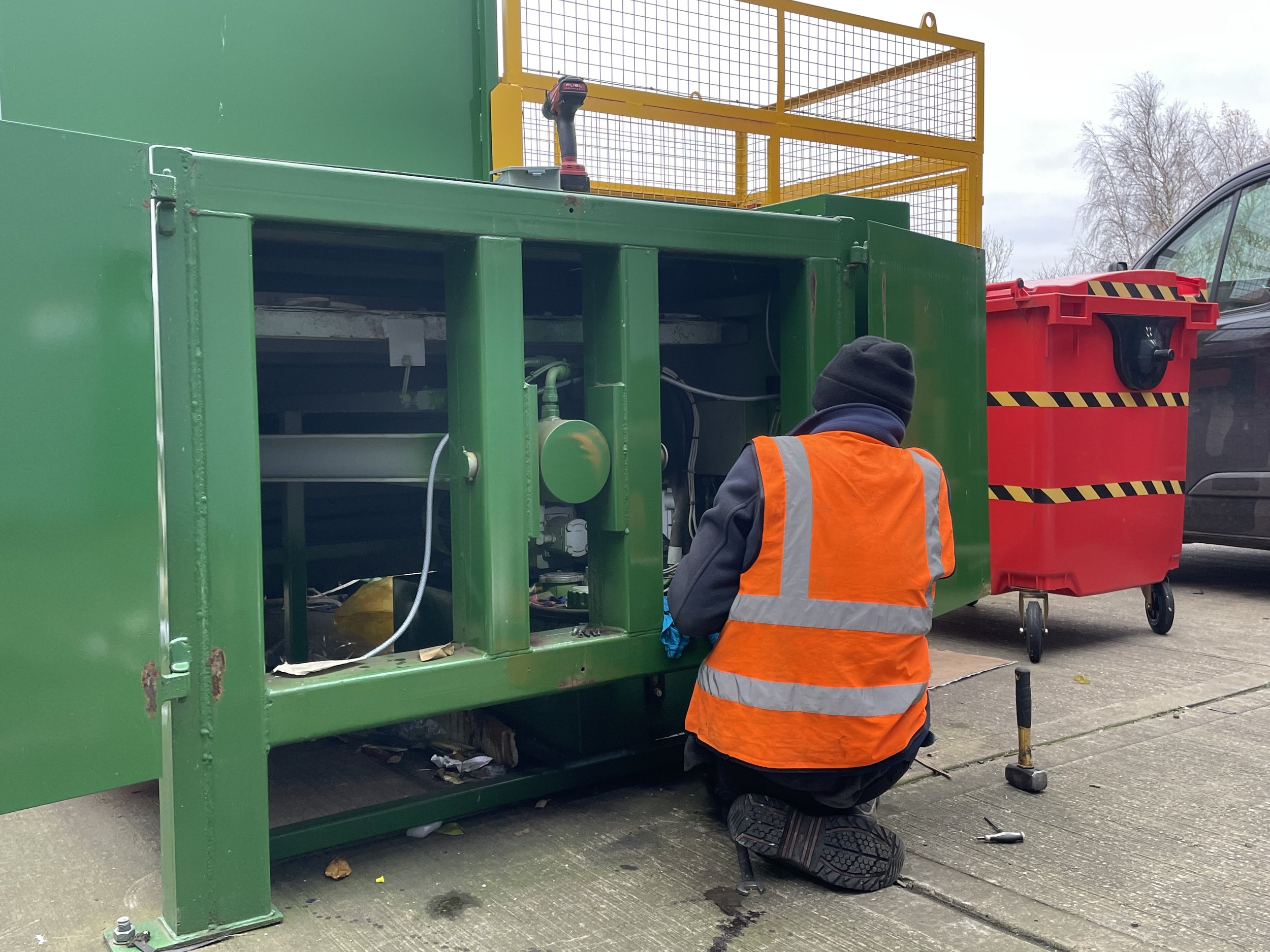 A maintenance worker wearing an orange safety vest and a black beanie kneels on the ground working on a large green industrial machine outdoors, with a red trash bin and a black vehicle nearby.