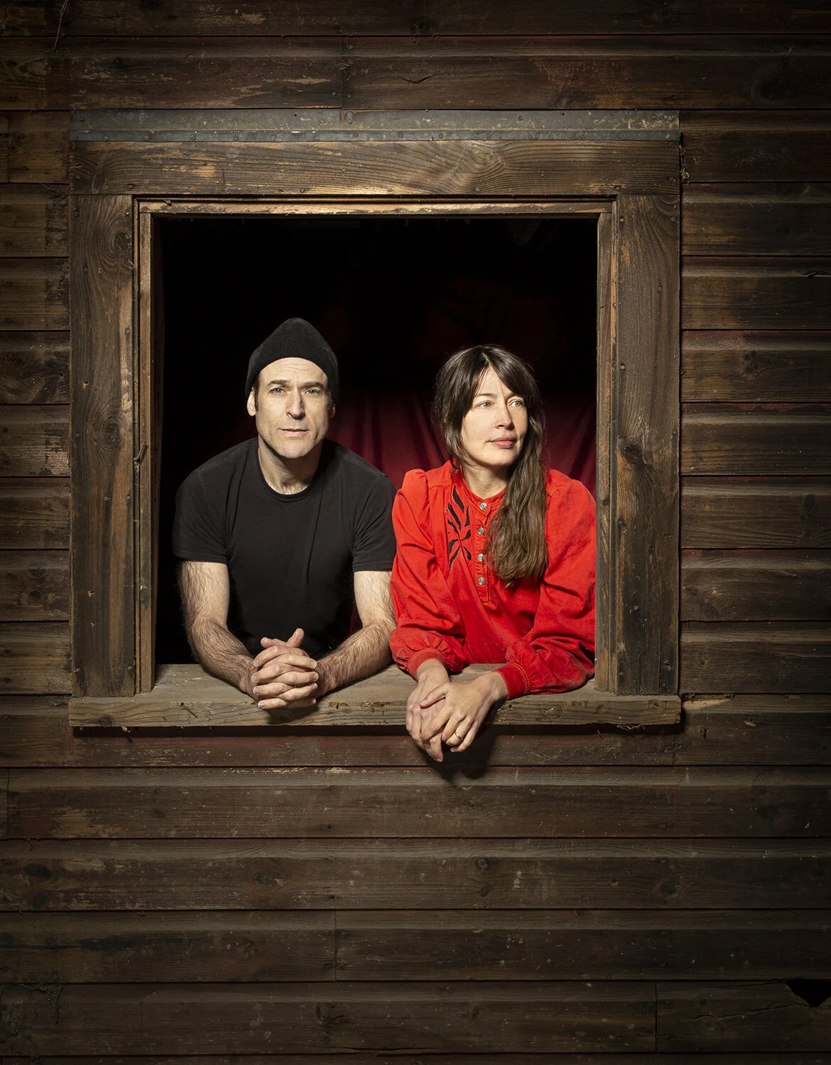 Alex Theodoropulos and Tara Foley leaning out of a wooden window frame, he in a black beanie and shirt, she in a red blouse