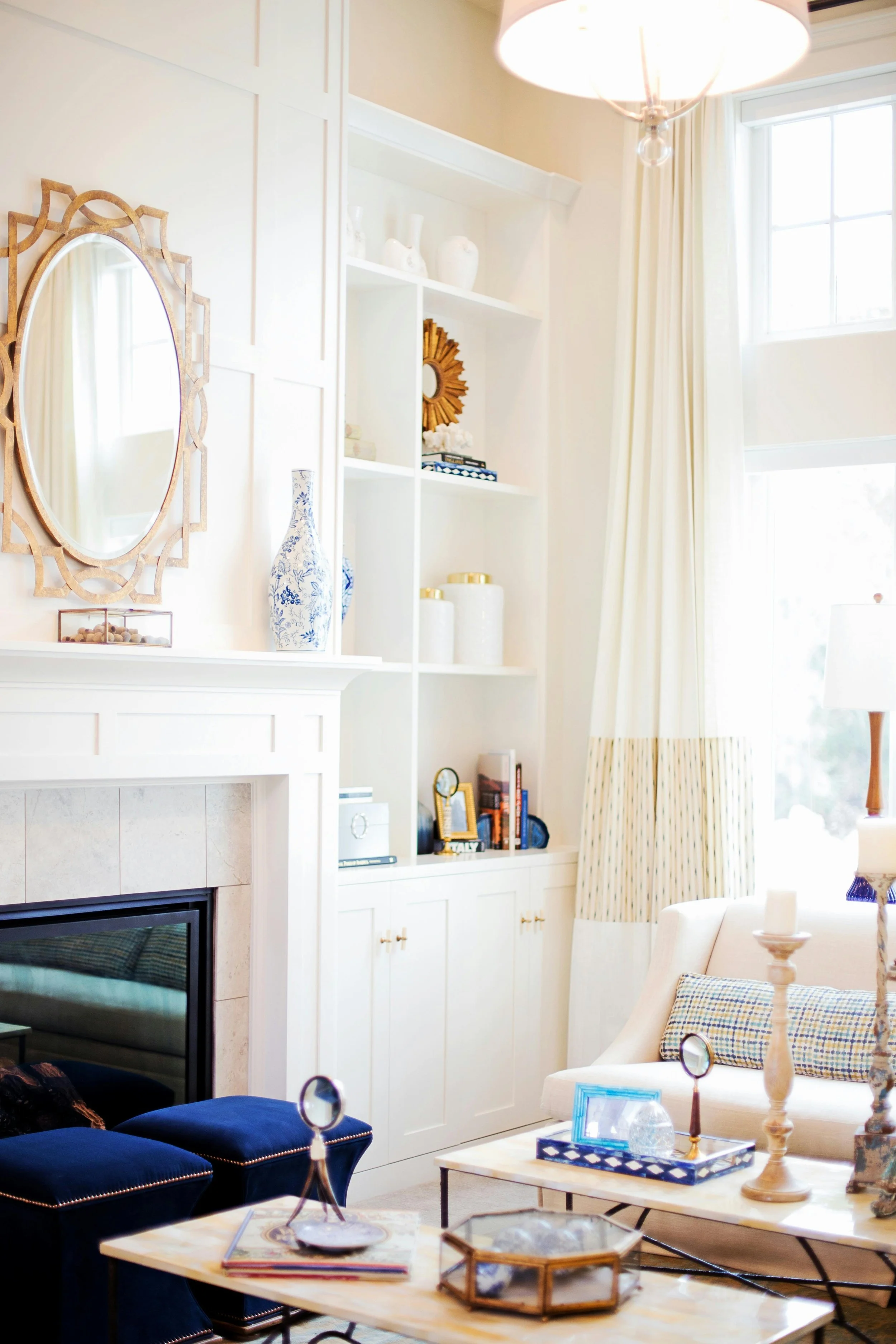 A well-lit living room with a fireplace, white built-in shelving with decorations, a large window with curtains, and a seating area including a white sofa, blue velvet ottomans, and a wooden coffee table with decorative items.